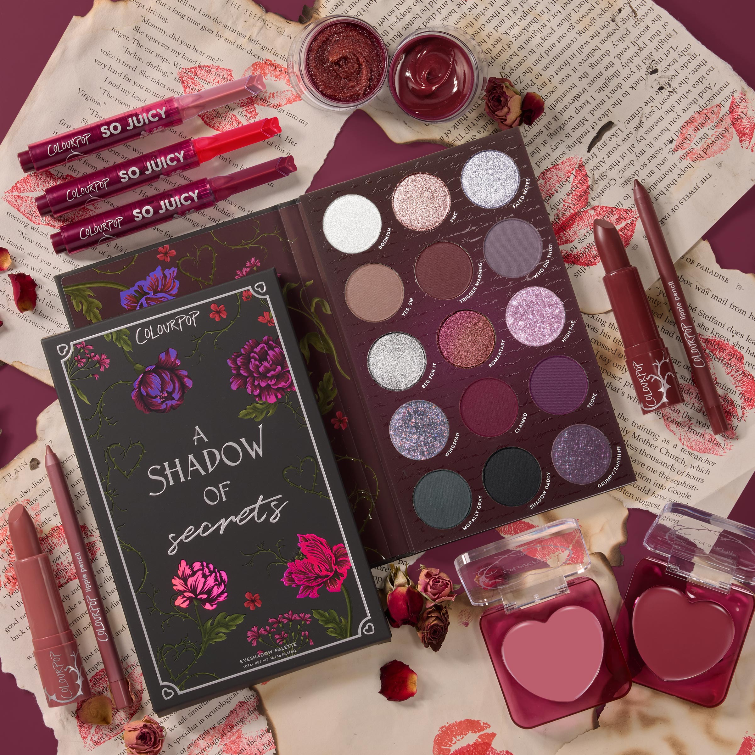 ColourPop Full Collection Set A Shadow of Secrets Collection arranged in a top-down flat lay, featuring a floral box, berry-toned eyeshadow palette, heart-shaped blushes, So Juicy glosses, lipsticks, liners, and cream pots on torn pages with rose petals.