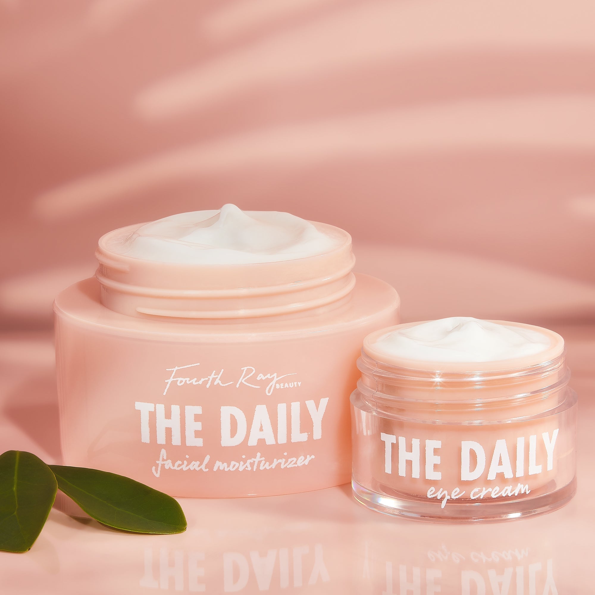 Fourth Ray Beauty The Daily Face + Eye Cream duo, jars opened to show whipped moisturizer textures, sitting side by side on a glossy pink backdrop with green leaves, a ColourPop Face & Eye Moisturizer Set for daily hydration.
