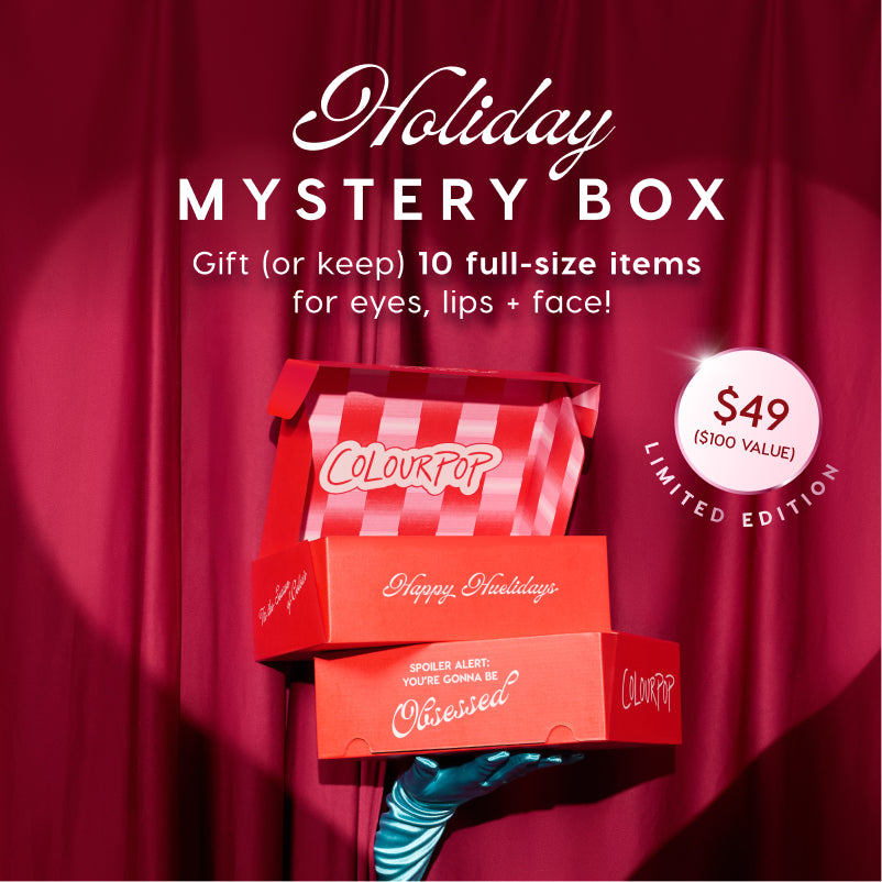 Colourpop Hauliday Mystery Box shown as stacked red mystery boxes, one open with red‑and‑white stripes, held by a teal gloved hand against red curtains; limited edition holiday set with 10 full‑size makeup items for eyes, lips, and face for $49.