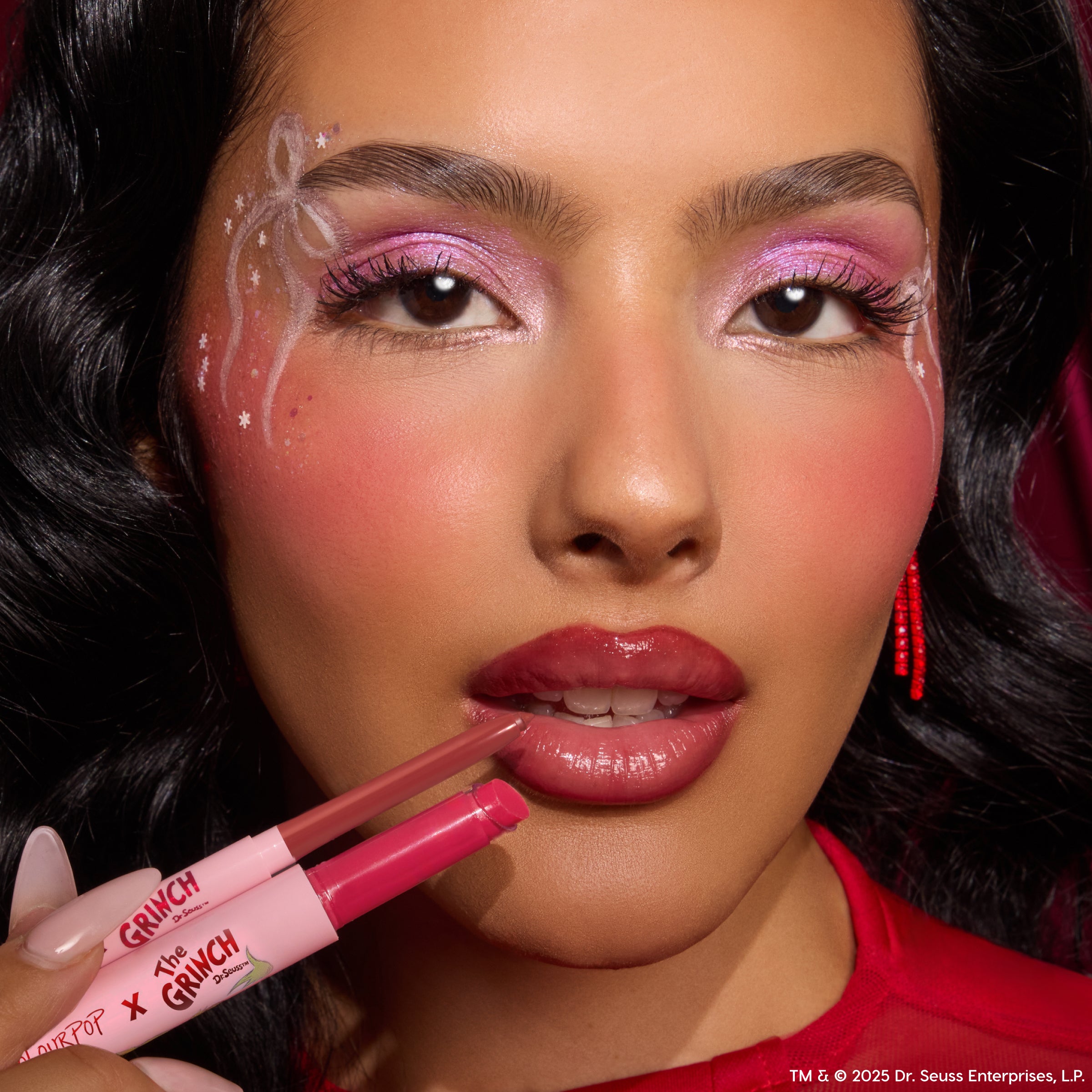 ColourPop So Juicy Plumping Gloss Stick in Berry Grinchy being applied to lips, delivering a plumped high-shine berry finish; close-up with pink shimmer eyeshadow, white ribbon face art, and red accents in a festive studio setting.