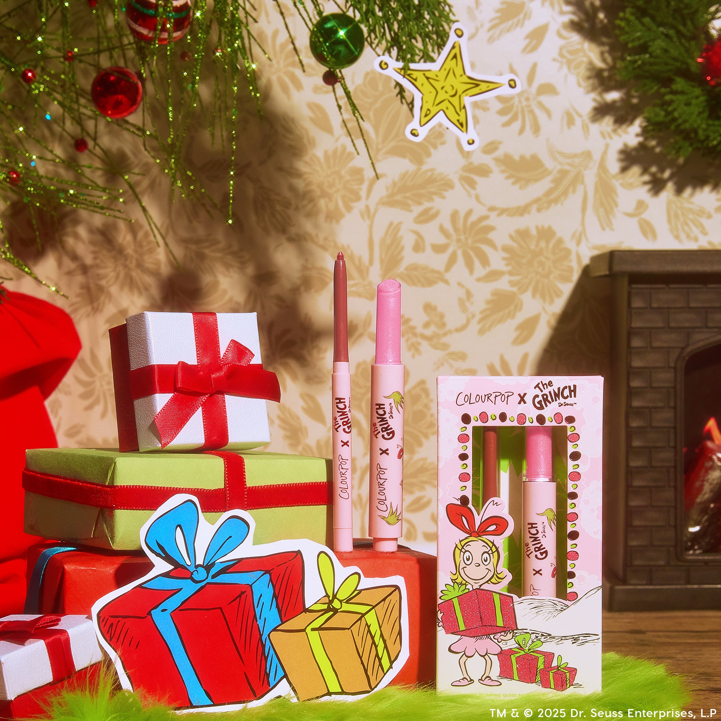 Pinky Who So Juicy Plumping Gloss Stick by ColourPop standing uncapped beside its boxed set, revealing a shimmery pink tip, arranged among wrapped gifts and ornaments; ColourPop x The Grinch packaging features Cindy-Lou Who art.