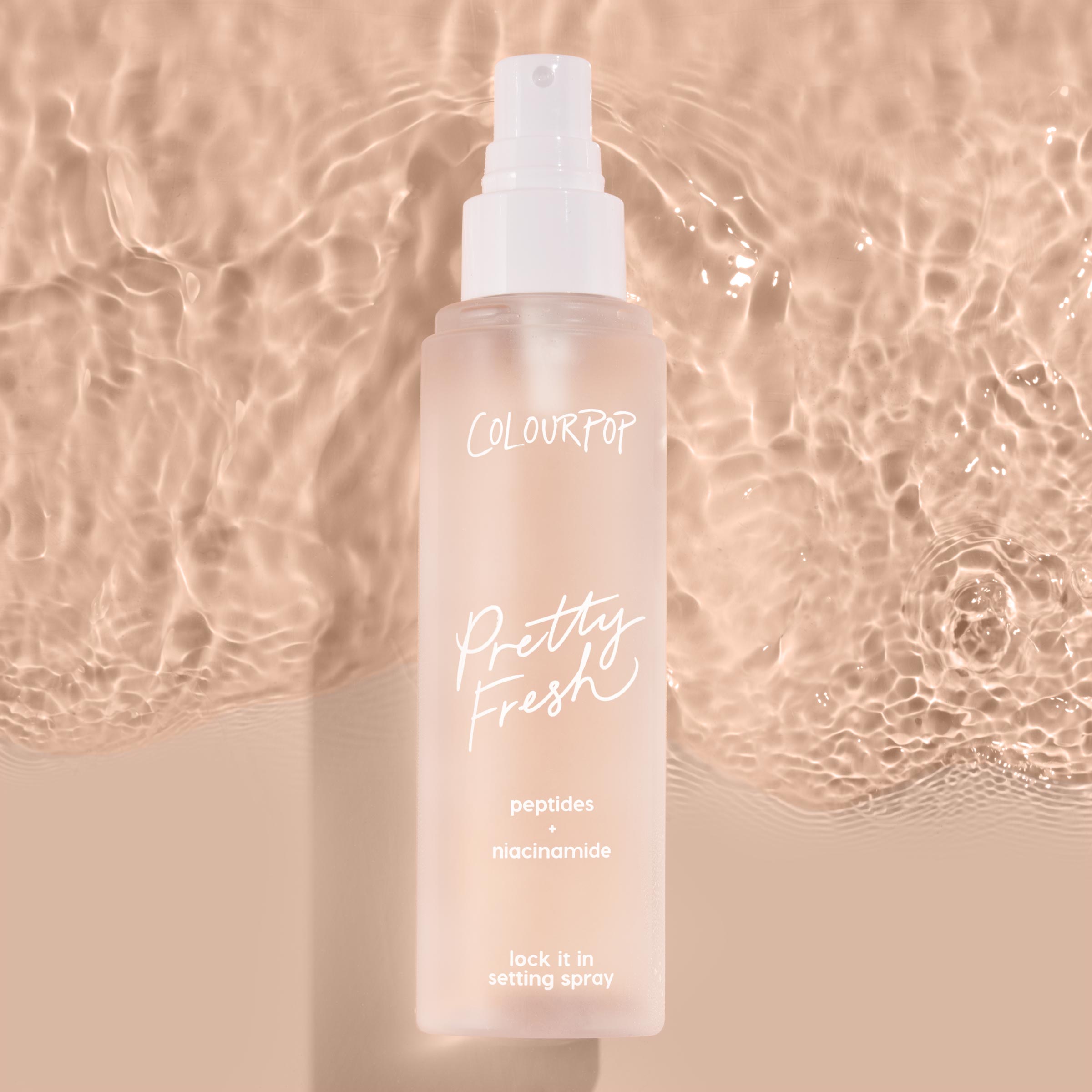 ColourPop Pretty Fresh Setting Spray, Lock It In, stands upright with a white pump on a frosted blush bottle labeled peptides + niacinamide, casting a soft shadow against rippled beige water for a clean, dewy makeup-locking look.