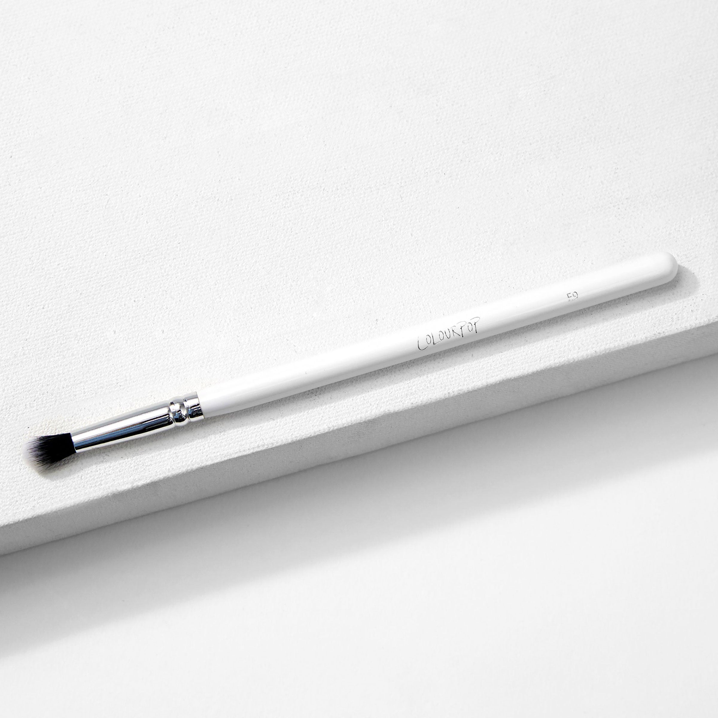 ColourPop Small Tapered Brush resting diagonally on a white textured platform, featuring a slim white handle, chrome ferrule, and small tapered black-and-white bristles for precise eyeshadow blending and detail work.