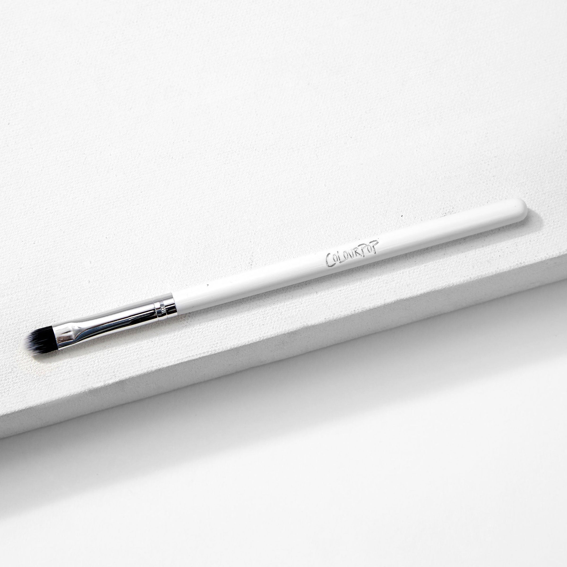 ColourPop Small Shader Brush makeup brush resting diagonally on a white textured surface, showing a sleek white handle with ColourPop logo, shiny silver ferrule, and dense flat rounded bristles for precise eyeshadow application.