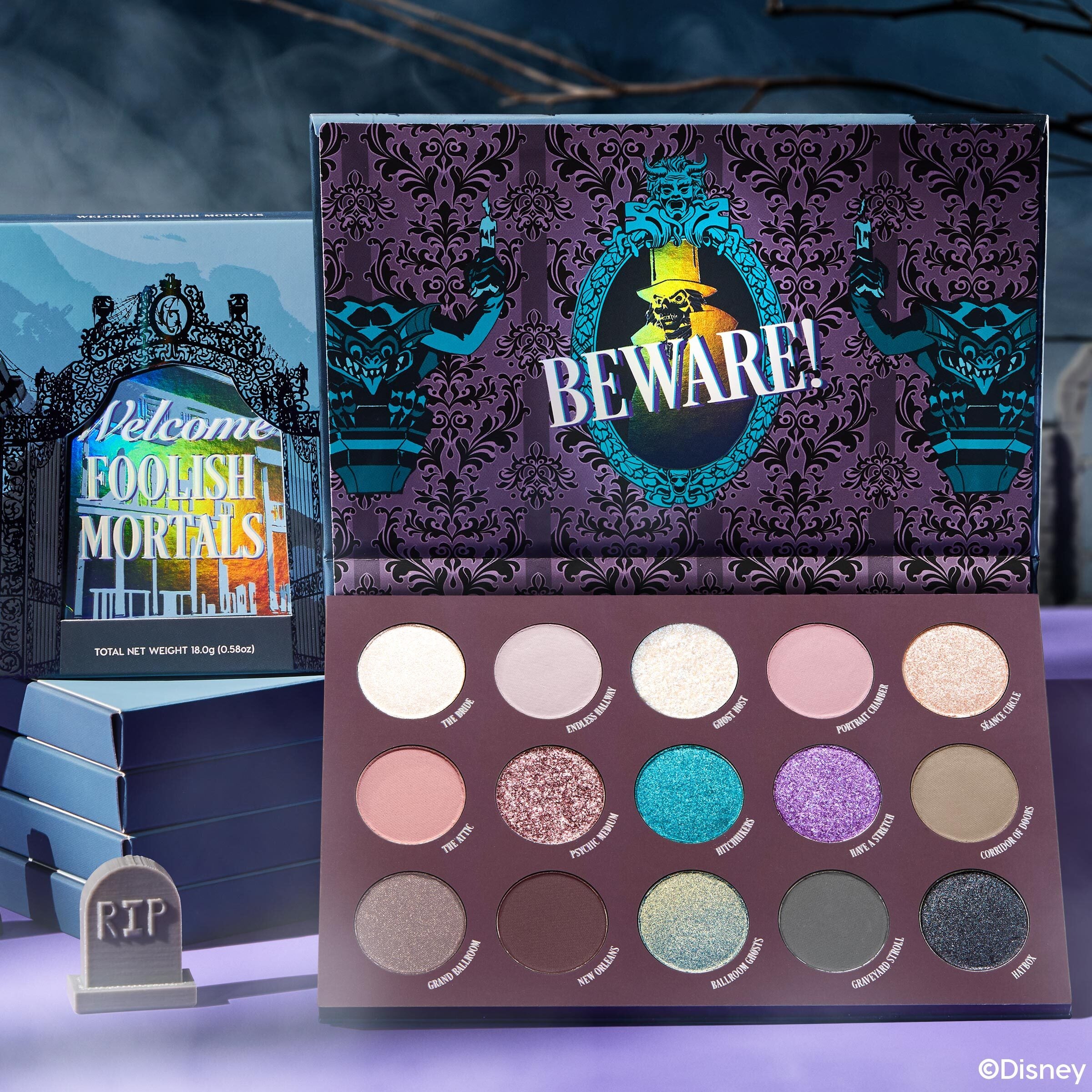 Welcome Foolish Mortals shadow palette by ColourPop opens to 16 haunted-mansion–inspired shades - champagne, taupe, teal, purple, black sparkle - in matte and shimmer finishes, shown with the 'Beware!' gothic interior and outer box on a foggy spooky set.