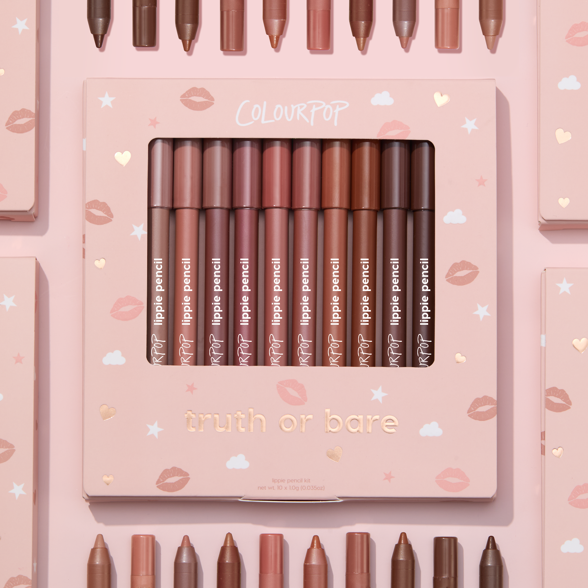 Truth or Bare Lippie Pencil kit by ColourPop displayed in a pink windowed box, 10 nude lip liner shades from peach to deep brown standing upright, surrounded by matching pencils and boxes on a pastel pink background with kiss and star icons.