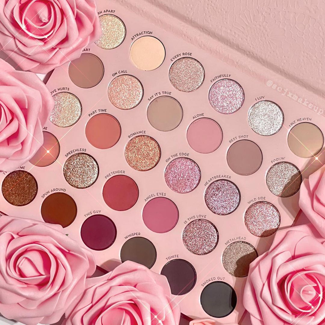 Smoke 'N Roses shadow palette by ColourPop lies open, displaying 30 rose-toned mattes, shimmers, and pressed glitters - from soft nude and mauve to deep charcoal and black - in a pink tray framed by pink roses; ColourPop Shadow Palette.