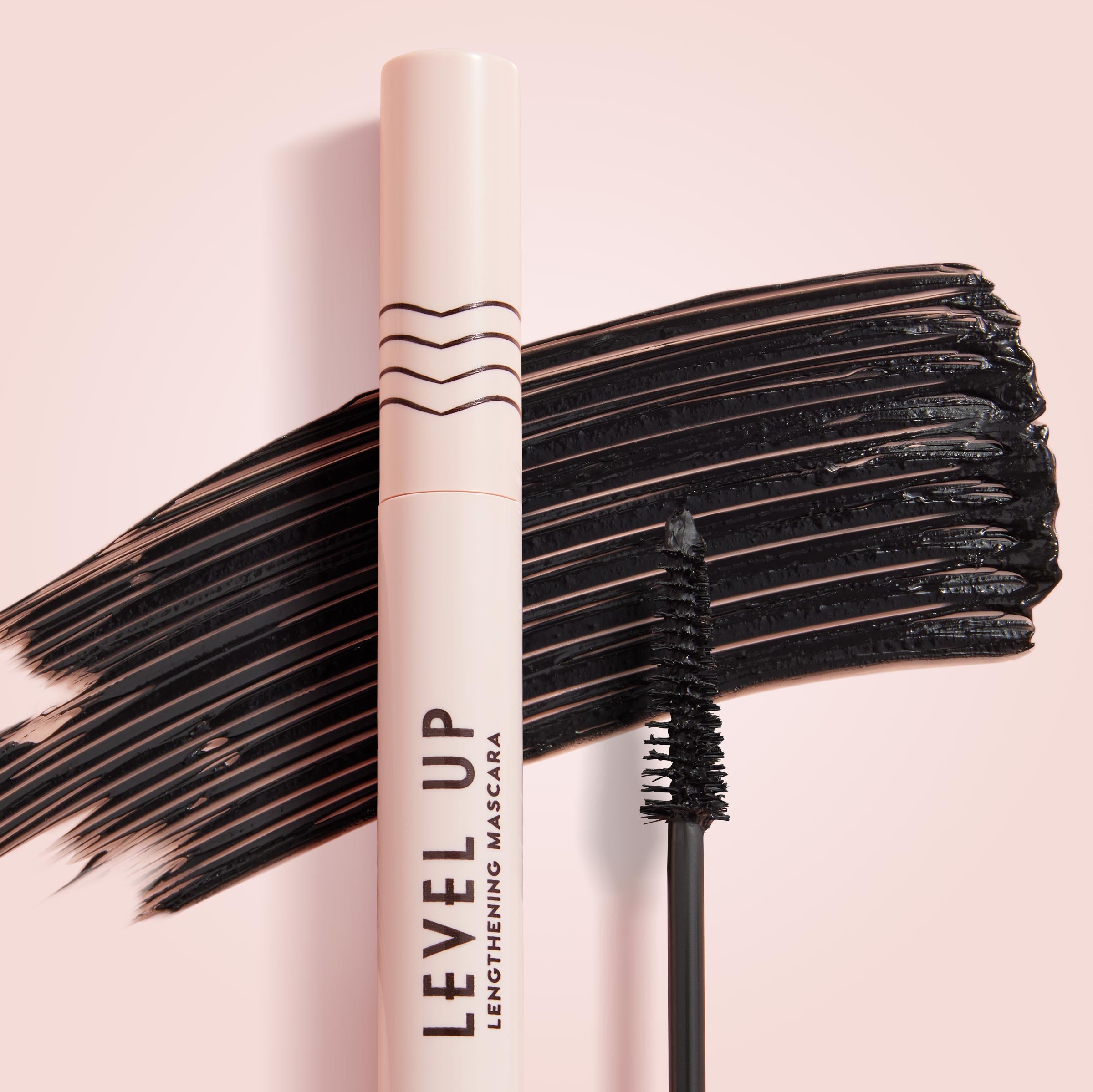 ColourPop Lengthening Mascara Level Up in Black stands upright with its wand coated in pigment, set before bold black swipes on a soft pink background, highlighting the slim brush and blush-pink tube for dramatic lash length.