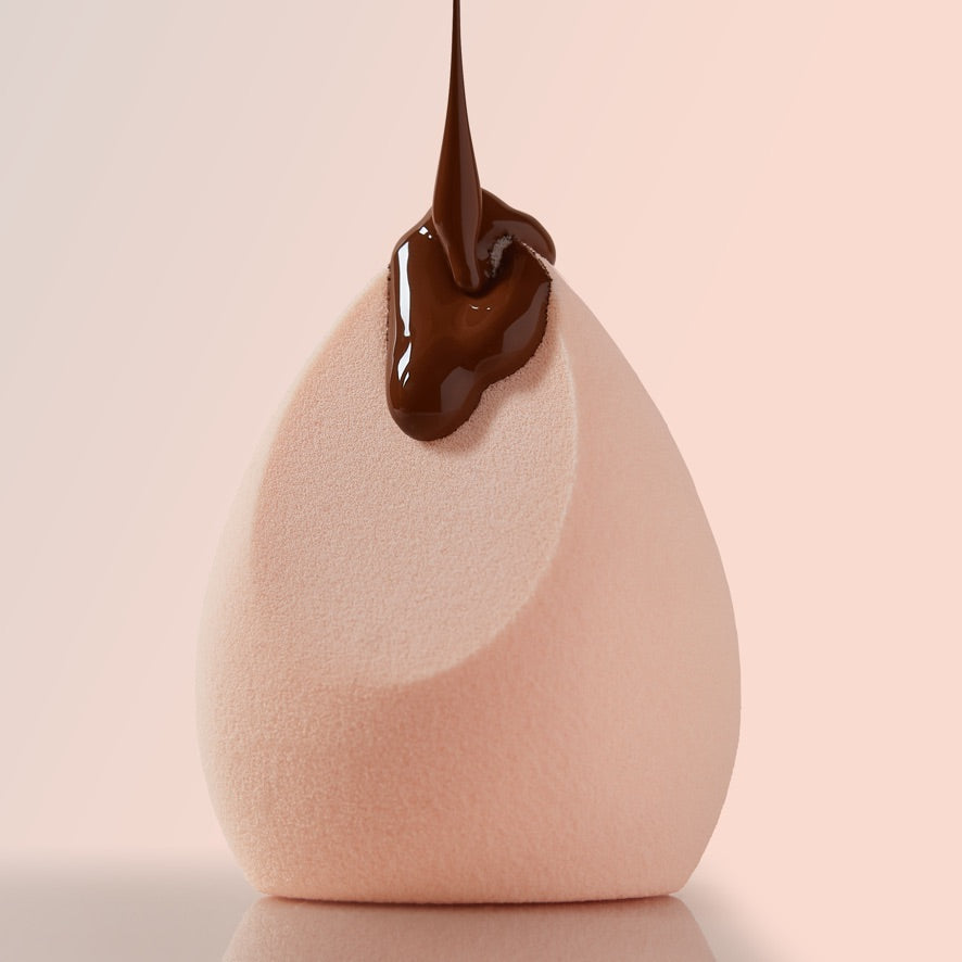 Pretty Fresh Complexion Blending Sponge catching a stream of chocolate-brown foundation on its slanted teardrop tip, shown close-up against a soft pink background; a ColourPop Makeup Tools & Accessories essential for seamless base application.
