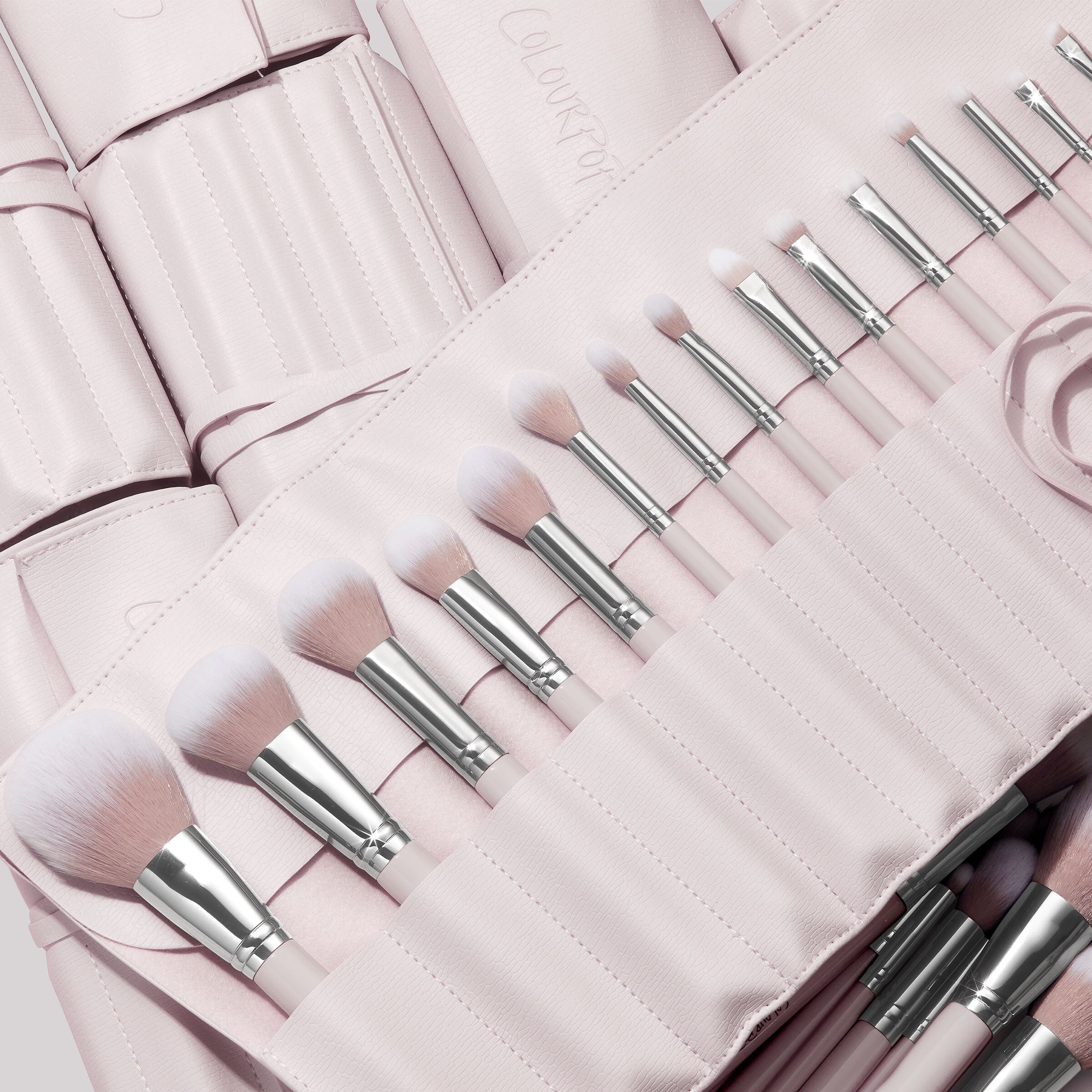 Stone Cold, a ColourPop Makeup Brush Kit, fanned in a blush-pink roll as face and eye brushes slot into stitched pockets, soft white-to-pink bristles and silver ferrules gleaming, with additional matching brush rolls layered behind.