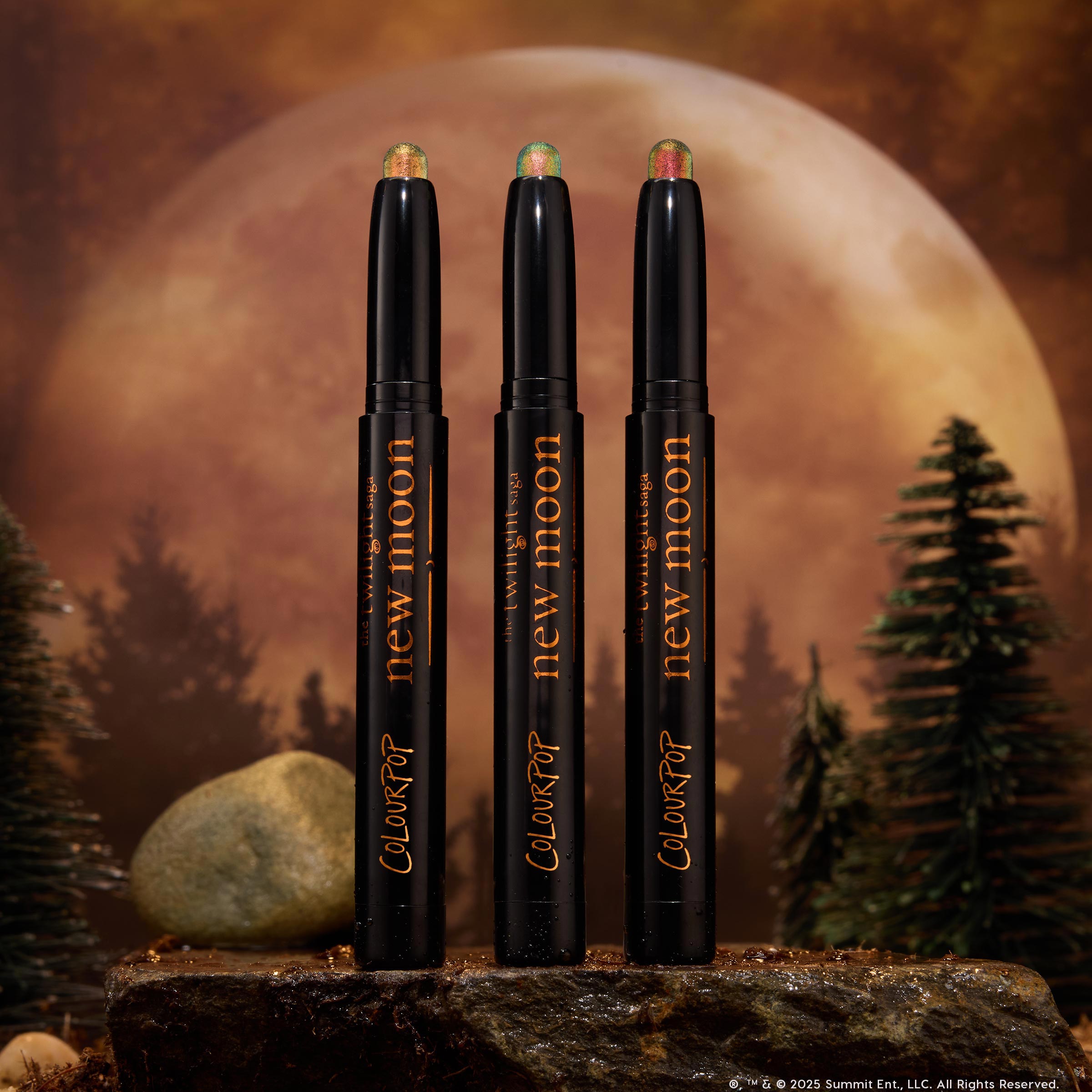 Three Love Triangle ColourPop Shadow Stix stand upright, their shimmery duochrome cream tips glowing green-gold and pink, displayed on a rocky surface with pine trees and a giant full moon backdrop for a moody, twilight woodland vibe.