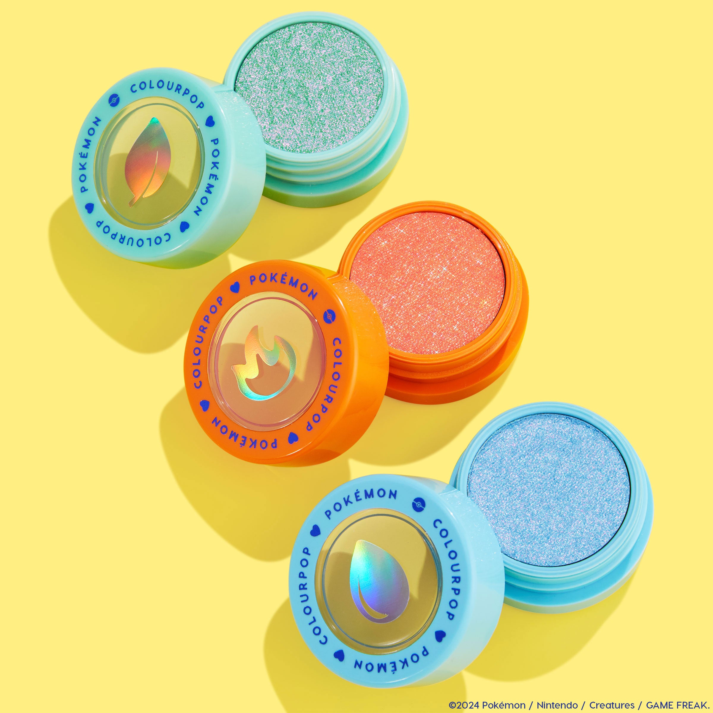 Flamethrower ColourPop Super Shock Shadow displays a sparkling coral-orange pan in an orange pot with a holographic flame lid, set on a bright yellow flat lay beside mint and sky-blue Pokémon-themed shadows.