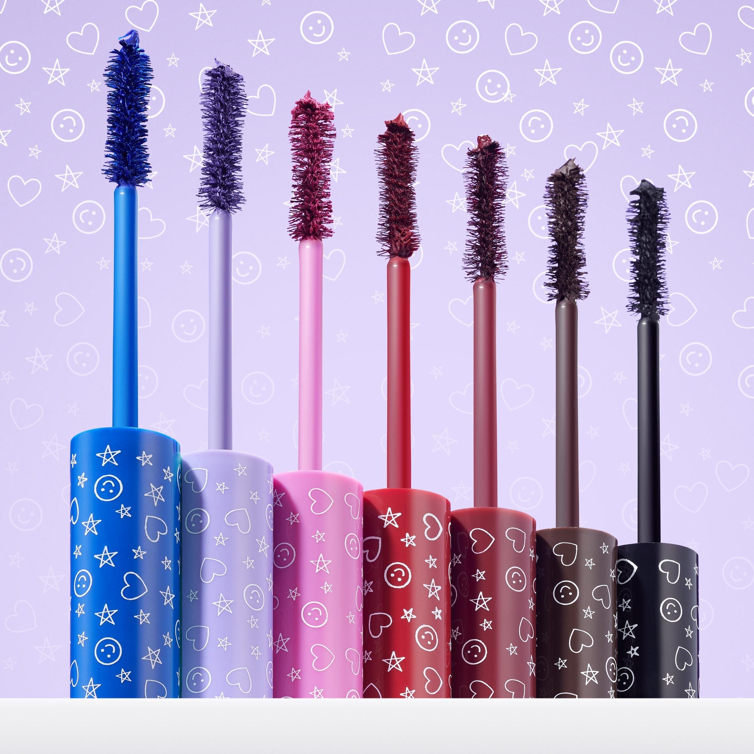 Slays of the Week ColourPop Mascara Set lined up, seven mascara wands - cobalt blue, lavender, hot pink, cherry red, burgundy, brown, and black - standing in tubes printed with stars, hearts, and smiley faces against a lilac backdrop.