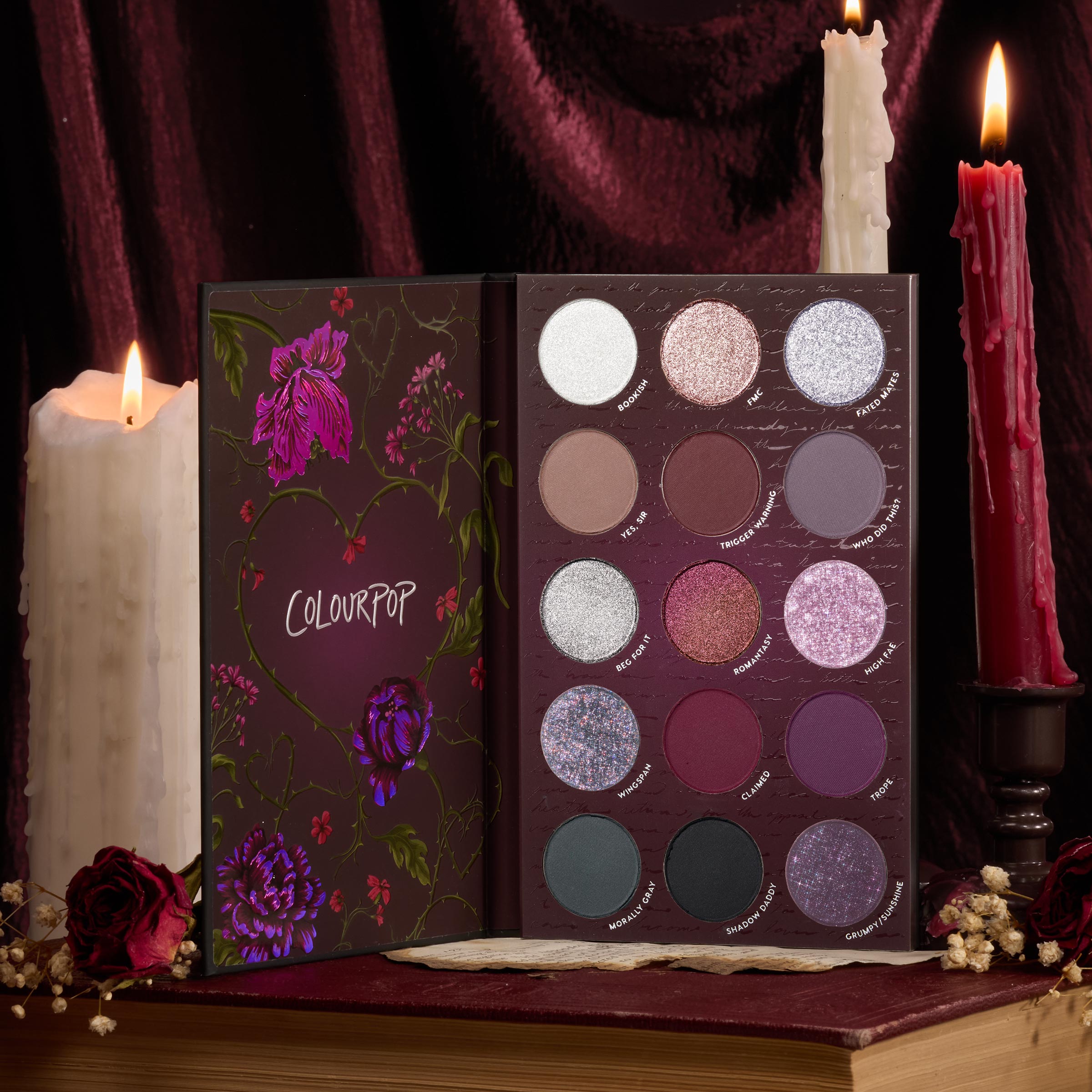 A Shadow of Secrets ColourPop Shadow Palette opens to 15 pans of cool-toned mauves, burgundies, and silver shimmers in a floral case, staged on a vintage book with lit candles and dried roses for a moody, velvet-draped setup.