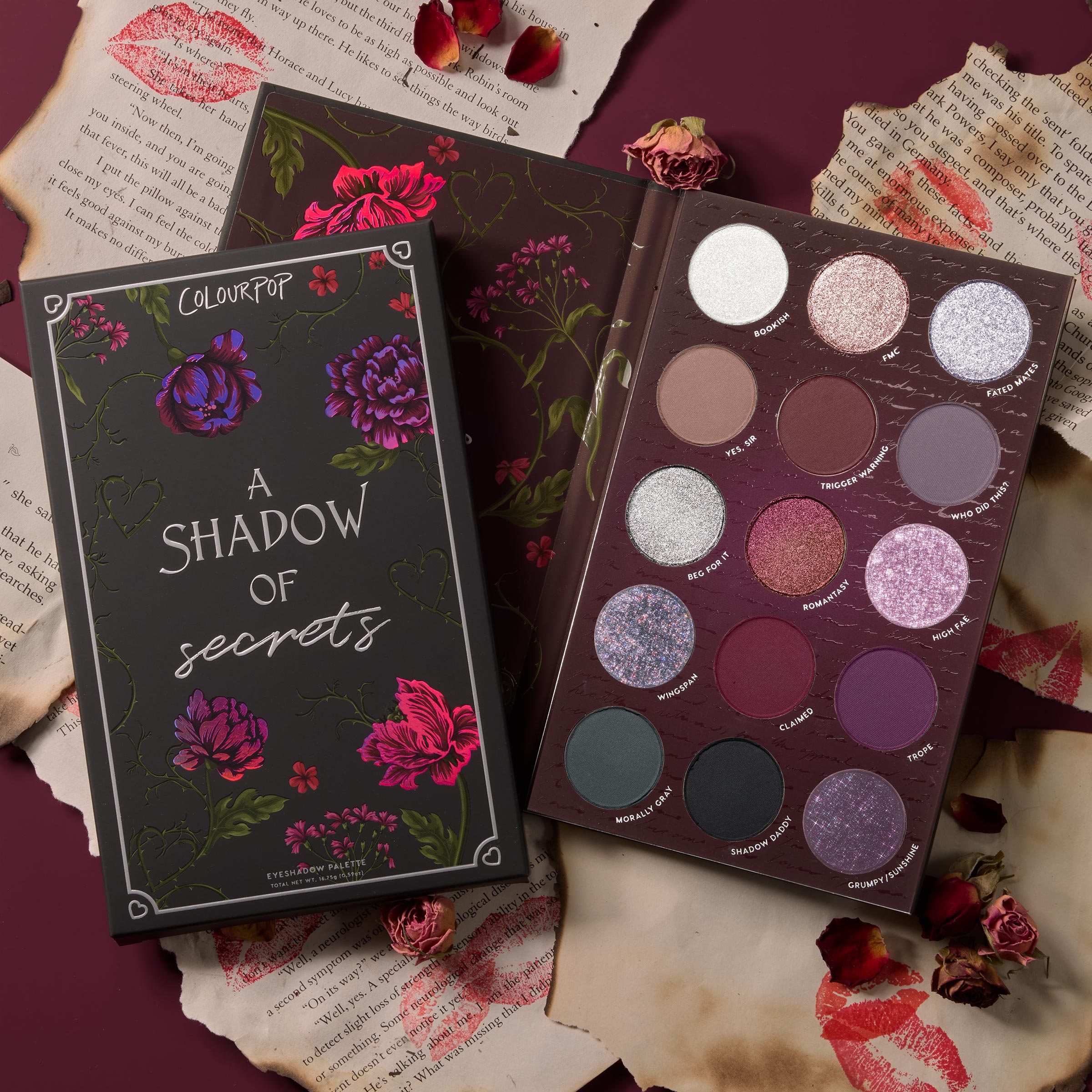 ColourPop A Shadow of Secrets shadow palette displays 15 shades - mauve, plum, silver, and charcoal - mix of mattes and sparkles, opened beside its floral box on a burgundy backdrop with torn pages, lipstick kisses, and dried roses; a romantic ColourPop Shadow Palette.