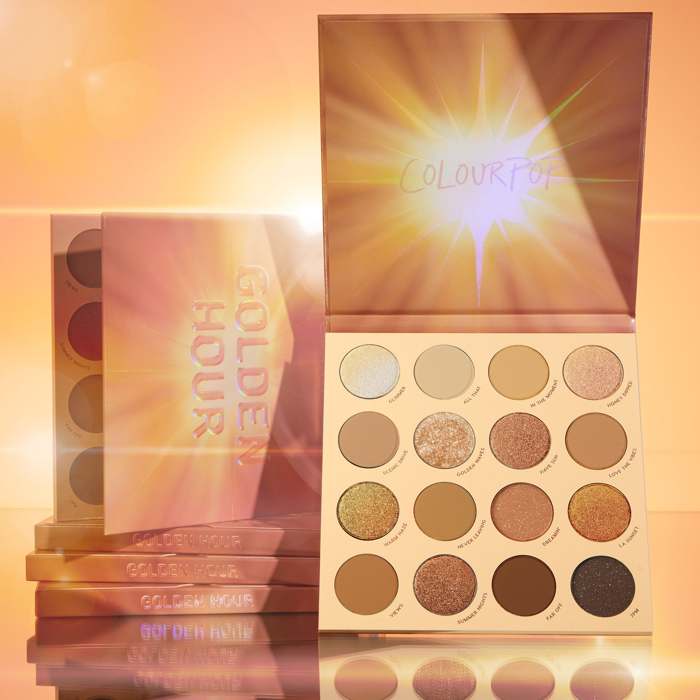 Golden Hour ColourPop Shadow Palette displaying a 16-pan mix of warm mattes, metallics, and glitter, lid with sunburst art propped open, with stacked palettes beside it on a reflective surface in a glowing golden studio.
