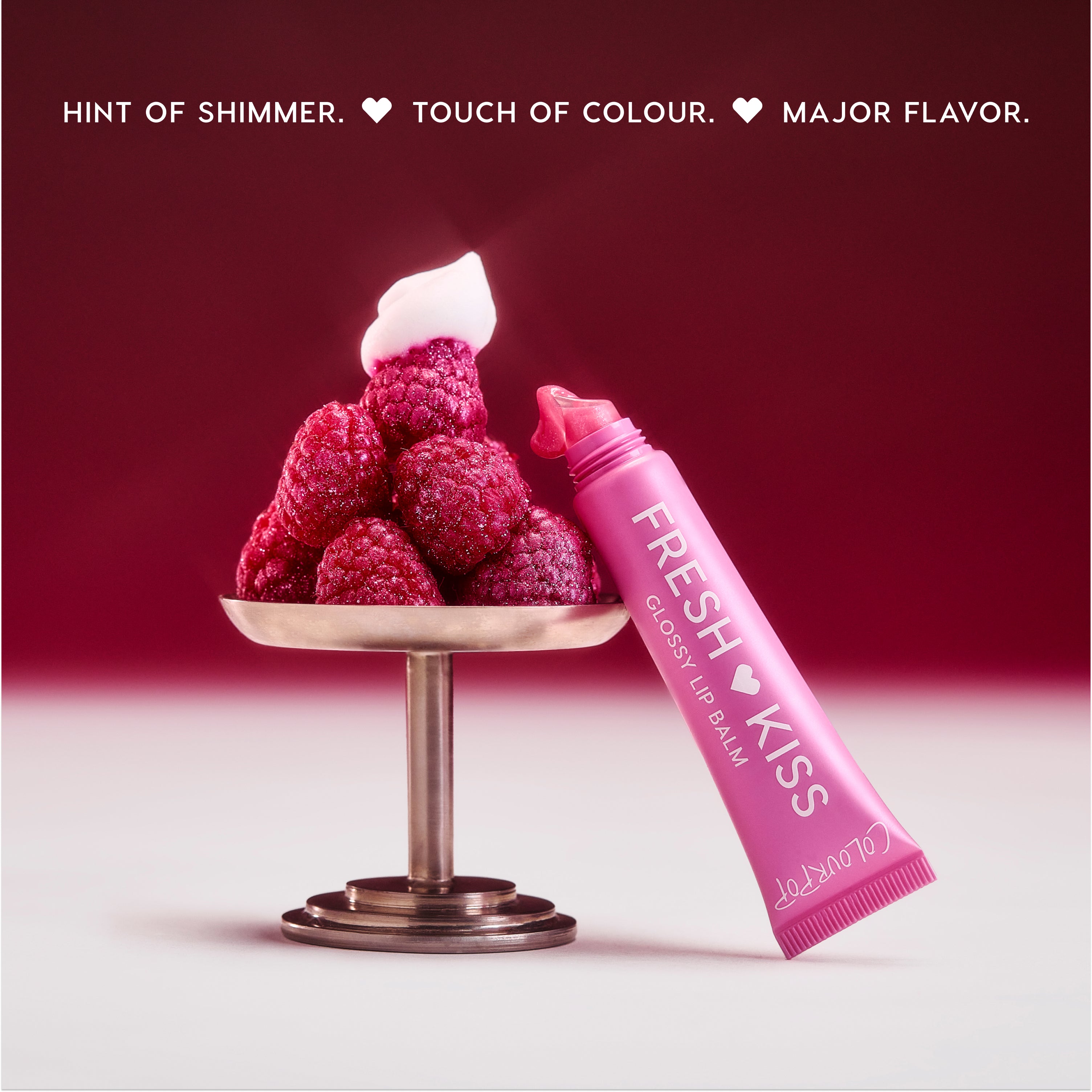 Colourpop Raspberry Crème Fresh Kiss Glossy Balm tube oozing pink shimmer next to a pedestal piled with fresh raspberries and whipped cream, highlighting juicy Lip Gloss & Lip Oil shine against a rich berry gradient background.