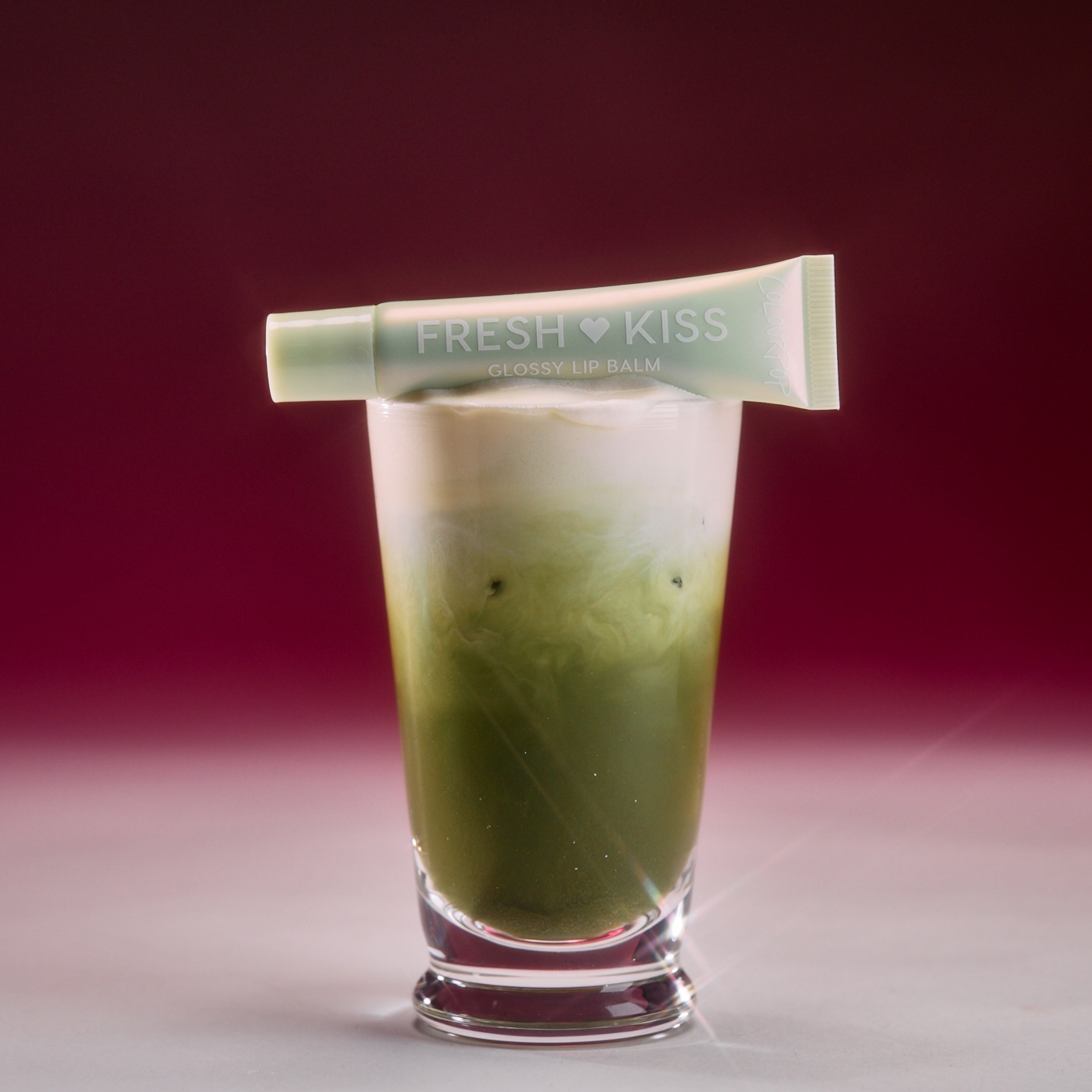 Colourpop Fresh Kiss Glossy Balm in Vanilla Matcha balanced across an iced layered matcha latte in a clear glass, highlighting the mint-green lip gloss & lip oil tube against a soft pink gradient background.