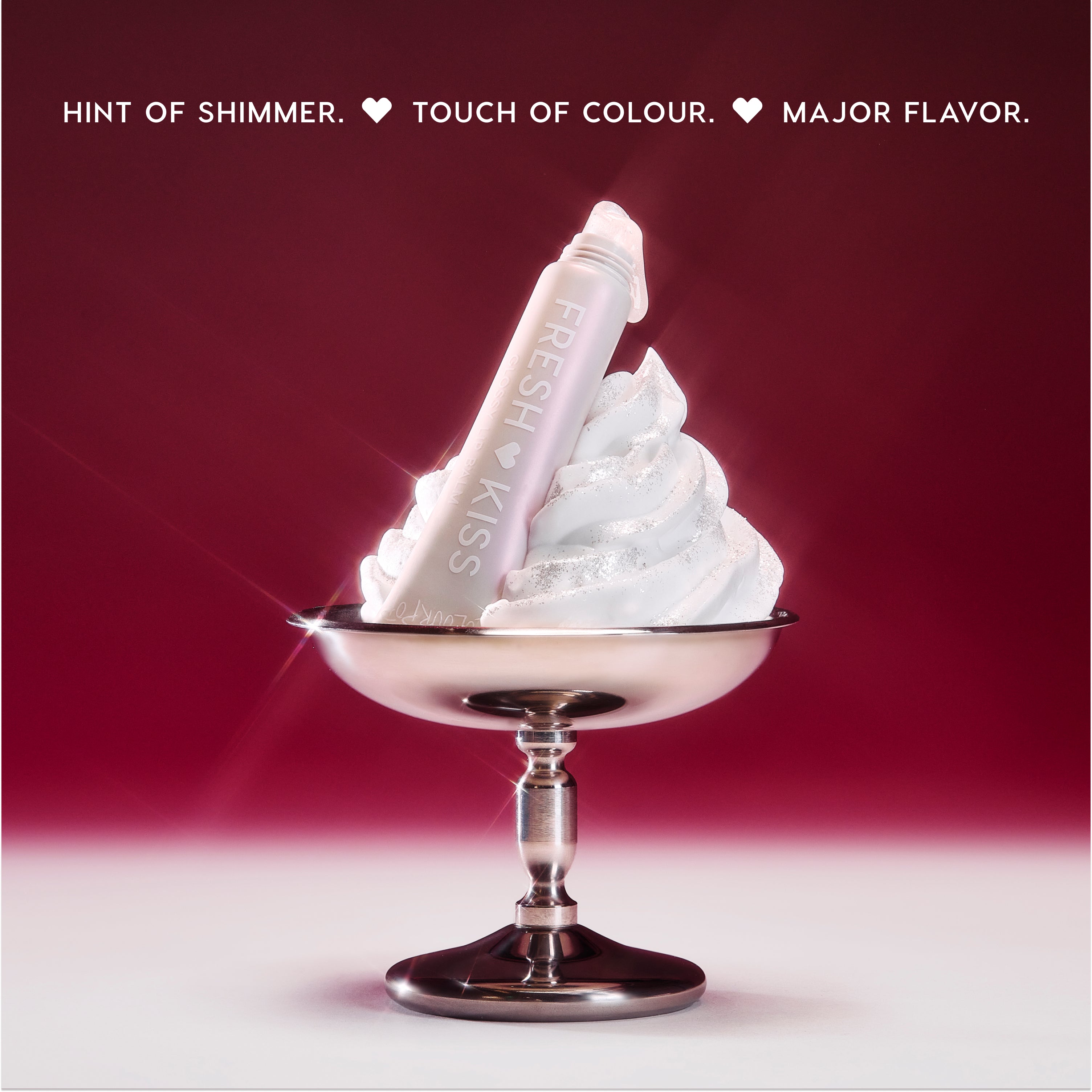 ColourPop Fresh Kiss Glossy Balm in Whipped Cream shade resting in a swirl of shimmering whipped cream in a silver dessert cup against a red gradient background, highlighting the glossy balm as a hybrid Lip Gloss & Lip Oil product.