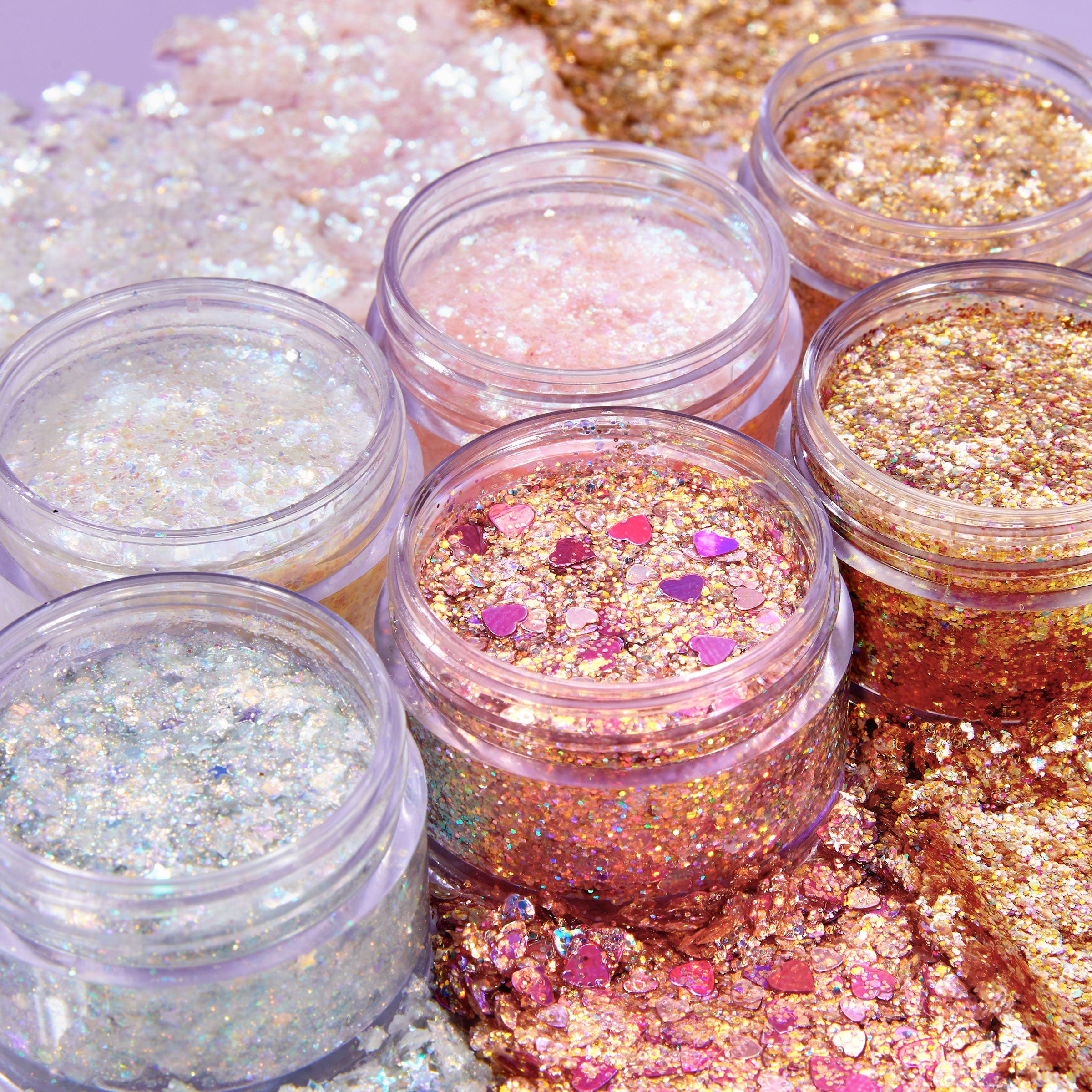 Ur A Firework ColourPop Glitter Gel jar gleaming with rose-gold glitter and fuchsia heart confetti, swatched beside other iridescent and gold glitter gel pots on a lavender surface to show the gel’s chunky texture and multi-dimensional sparkle.