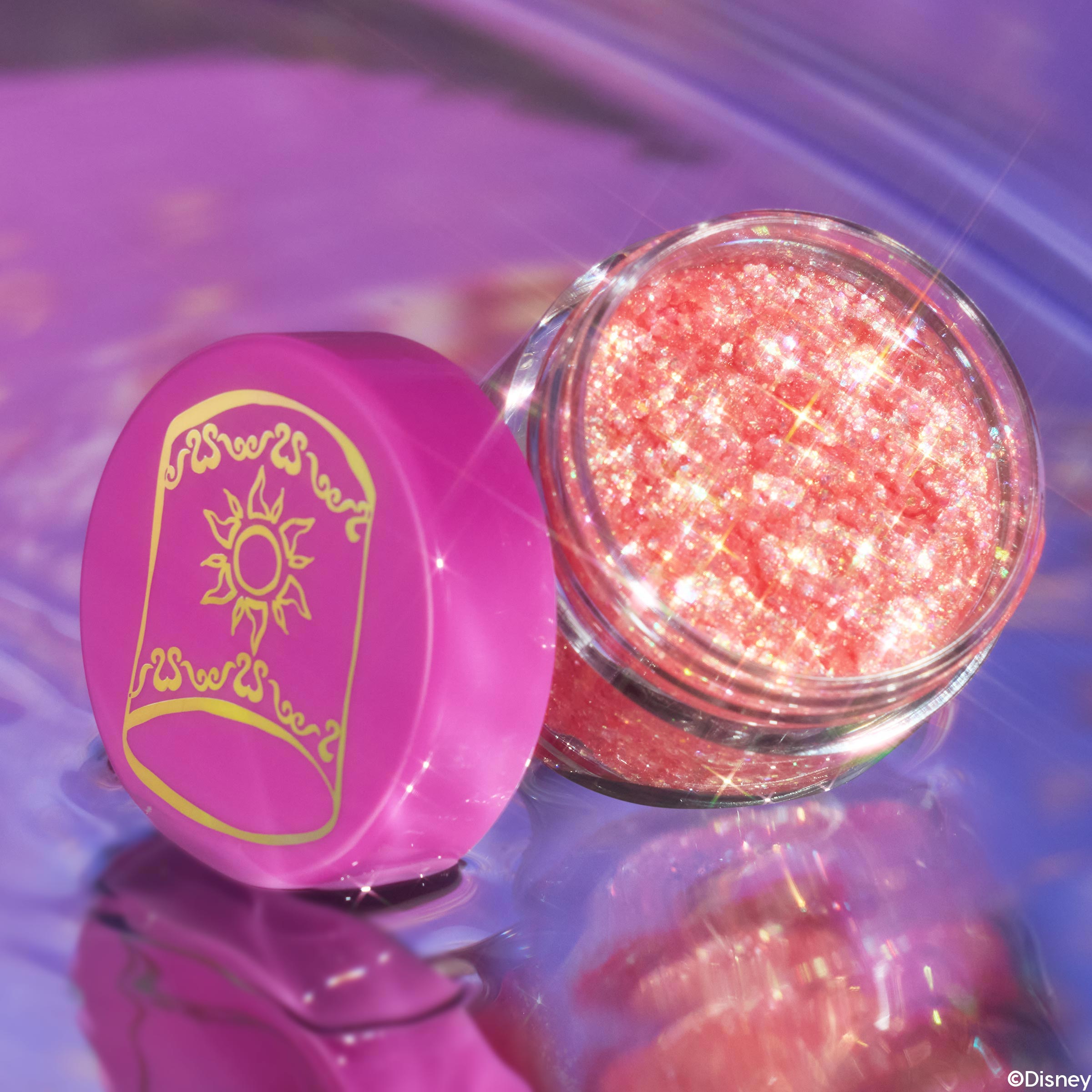 Open jar of ColourPop Glitter Gel, Floating Lights, gleams with peachy-pink iridescent flecks; the clear pot rests on a glossy lavender reflective surface with a magenta lid featuring a gold sun motif beside it.