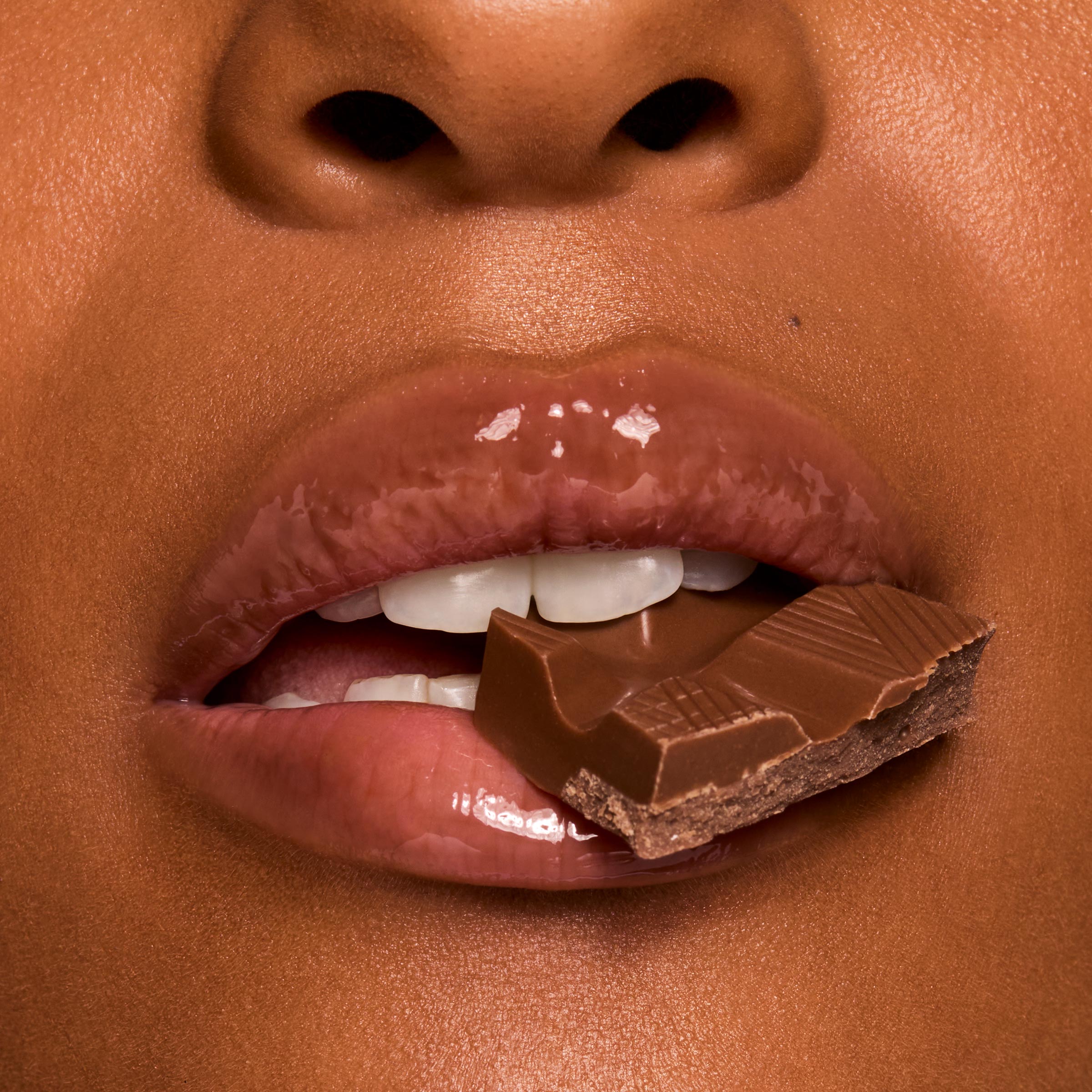 Glossy lips bite a piece of milk chocolate, showcasing rich shine in a chocolate-brown shade from Gloss Goals, a ColourPop Full Collection Set, in a tight beauty macro emphasizing a smooth, hydrated finish.