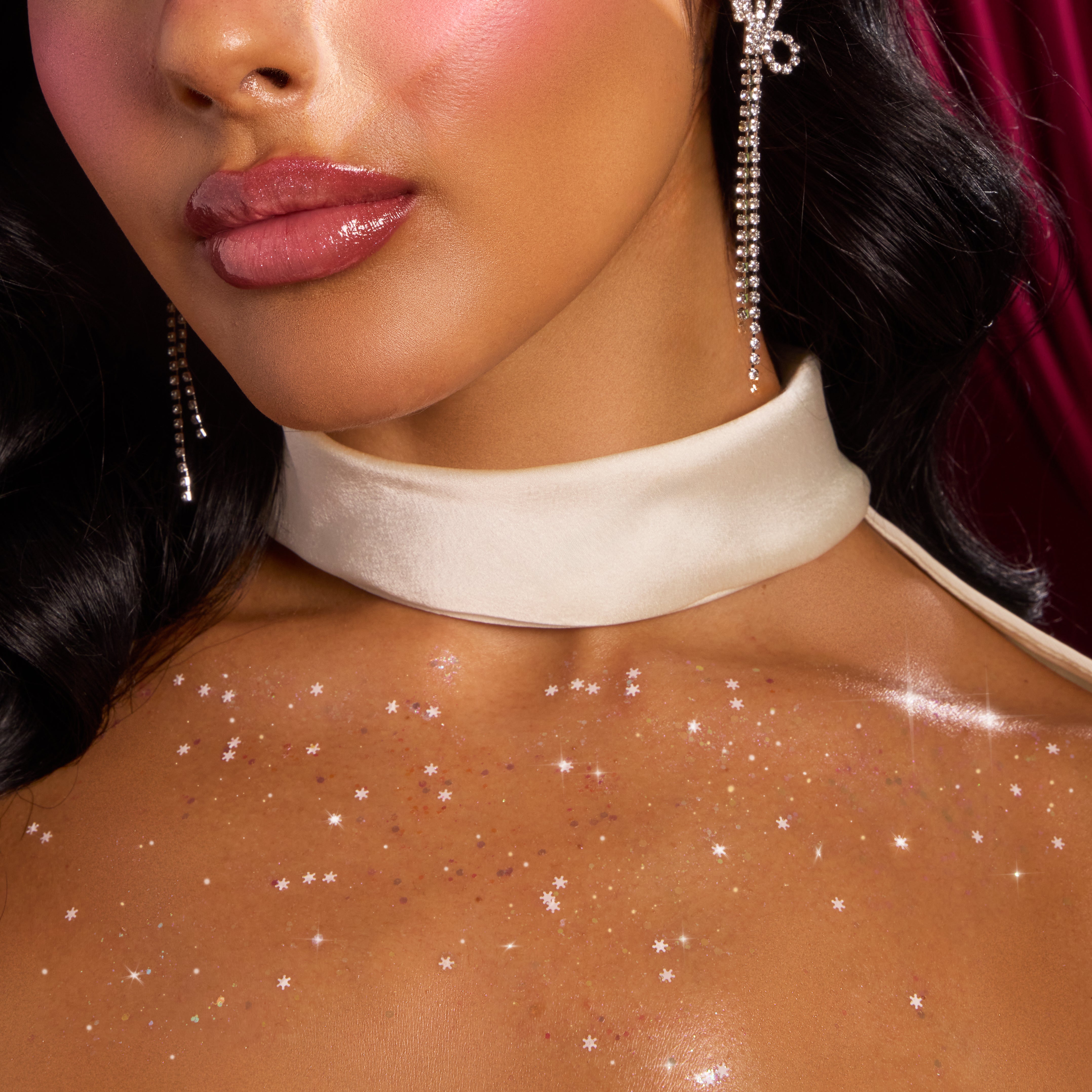 ColourPop Who-Hash Glitter Gel shimmering across a model’s collarbone and chest, scattering star-shaped sparkles; close-up with glossy lips, satin ivory choker, and rhinestone drop earrings against a deep red curtain backdrop.