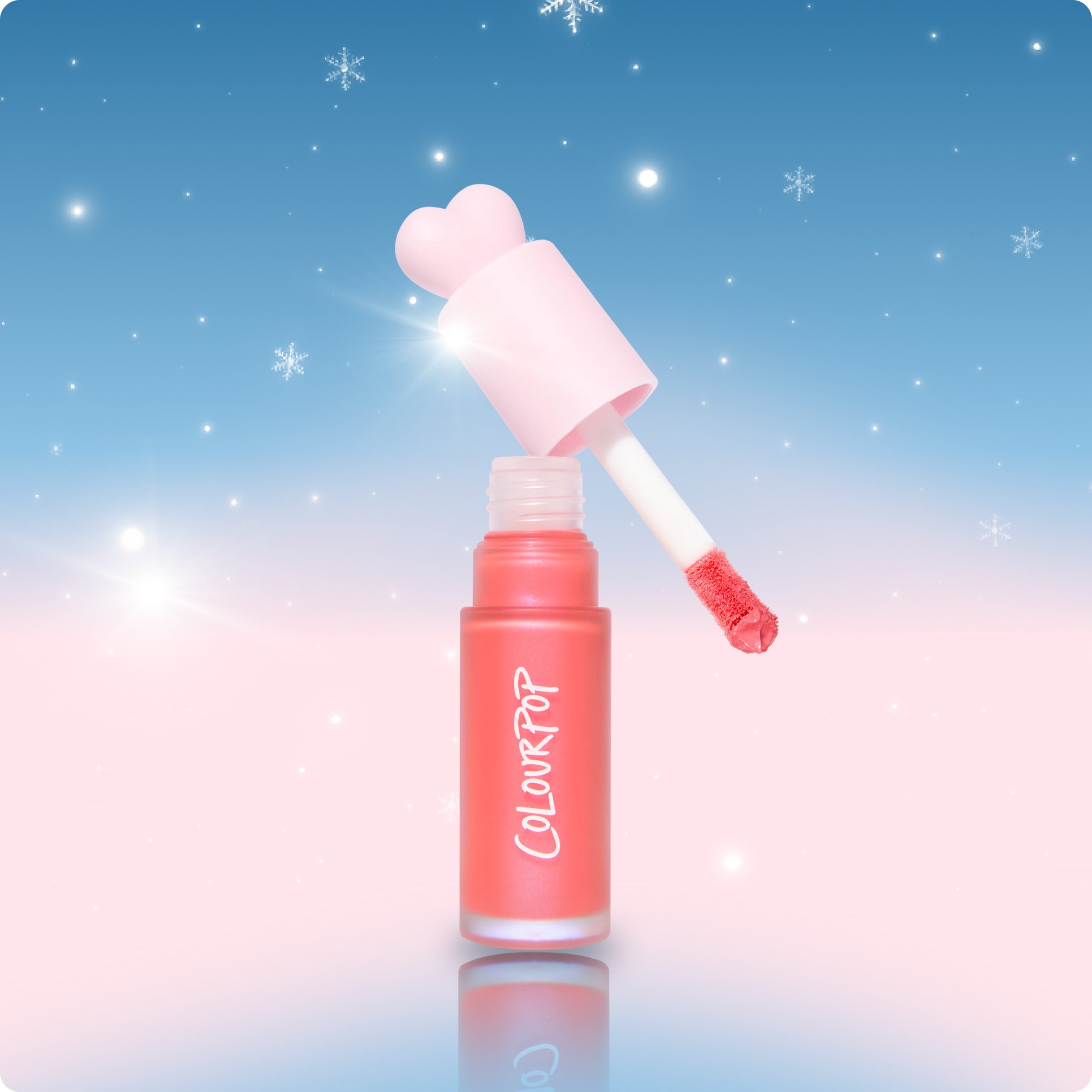 Colourpop pink liquid lip gloss with heart-shaped wand cap hovers open, showing coral applicator against a dreamy blue-to-pink gradient holiday background with sparkling snowflakes, highlighting festive US makeup gift vibes.