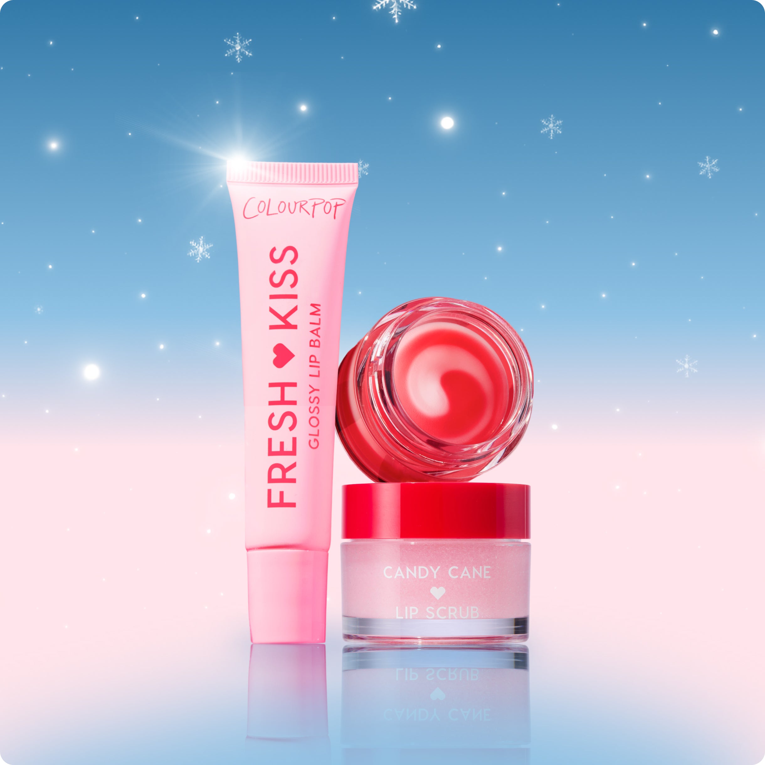 Colourpop Fresh Kiss glossy lip balm tube and Candy Cane lip scrub jar arranged upright on a reflective surface against a pastel blue and pink snowy holiday background, highlighting festive pink-red lip care set for smooth, glossy lips.