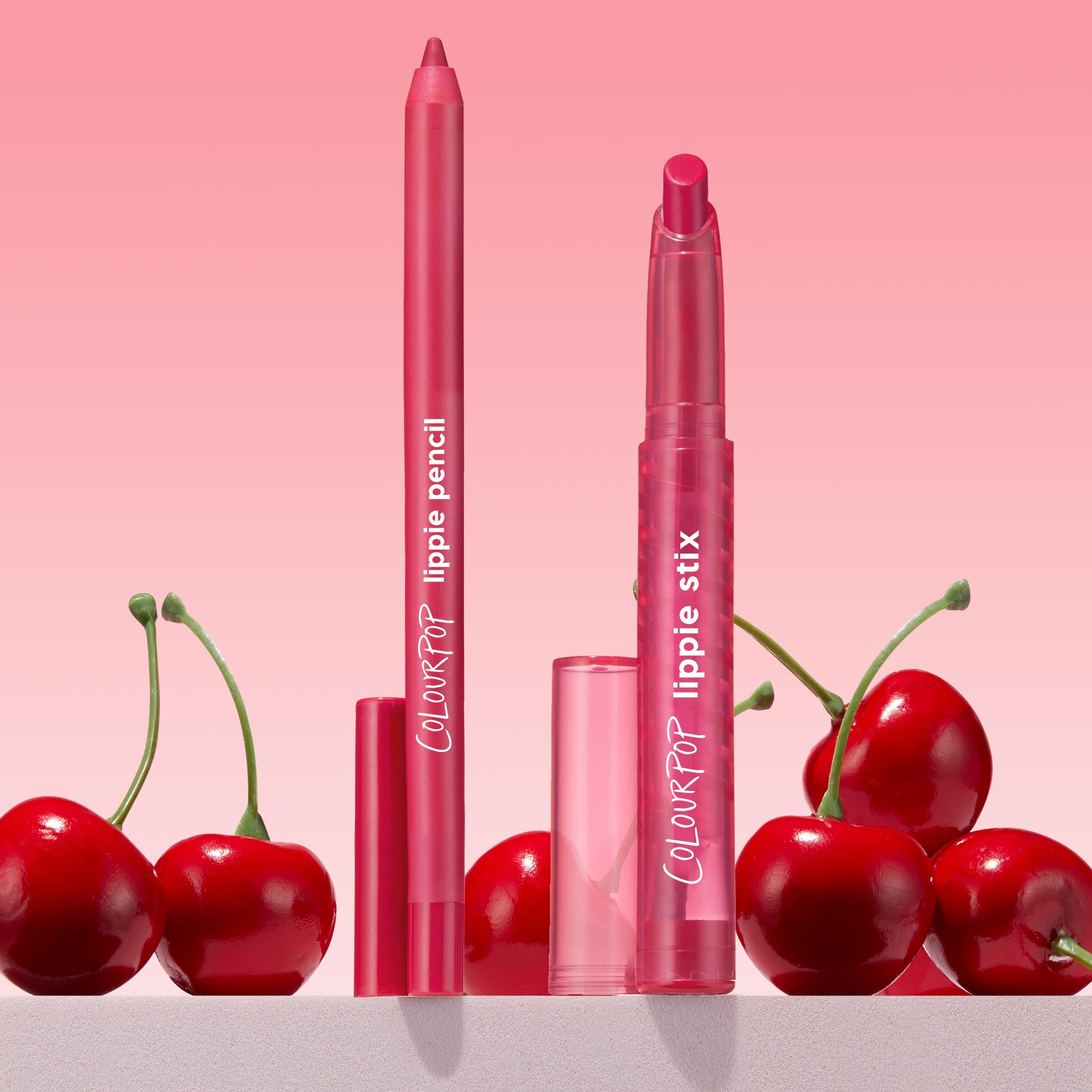 Ultimate Lip Combo showcases a pink-red ColourPop Lippie Stix + Pencil Set: lippie pencil and lippie stix standing upright with translucent pink caps, framed by fresh cherries against a soft pink gradient backdrop.