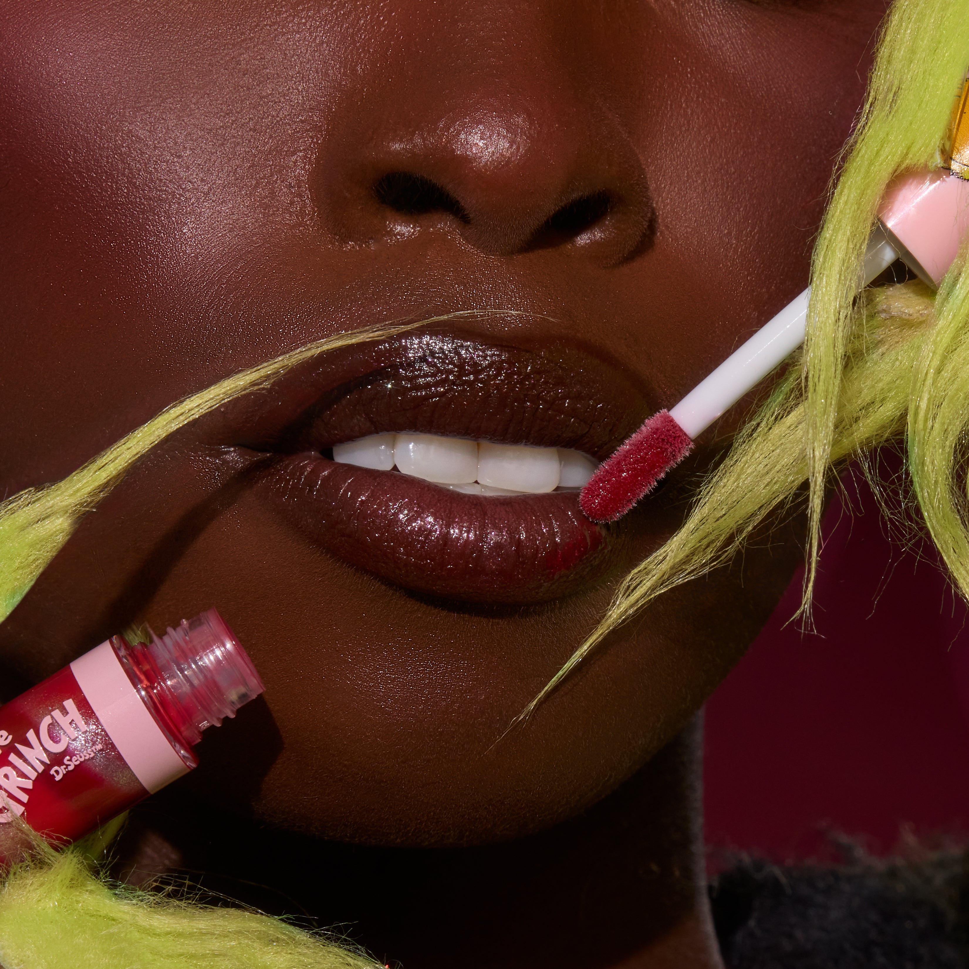 Glossy lips receive a swipe of ruby swirl lip gloss from a doe-foot applicator, with the open red tube nearby, highlighting the ColourPop Merry Grinchmas Swirl Lip Gloss Kit amid lime-green faux fur accents and a deep magenta backdrop.