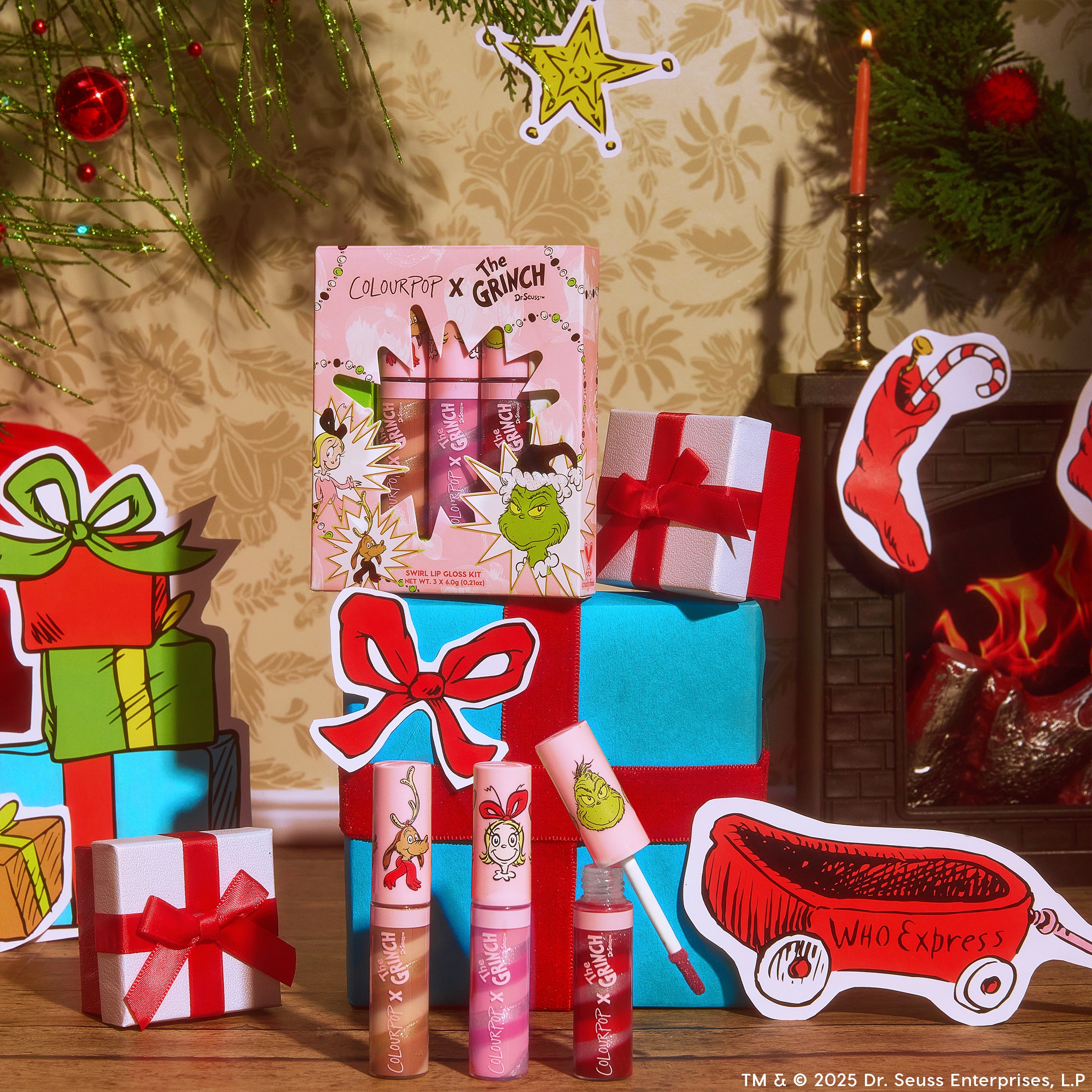 ColourPop Merry Grinchmas Swirl Lip Gloss Kit featuring a 3-piece set of pink-toned swirl lip glosses, one wand open, displayed in Grinch-themed packaging amid wrapped gifts and a lit fireplace for a festive holiday scene.