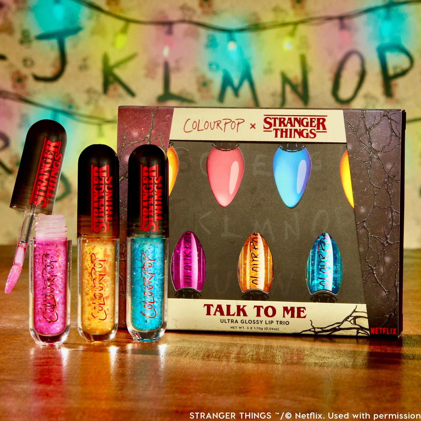 Stranger Things x ColourPop Full Collection set showcasing the Talk To Me ColourPop Ultra Glossy Lip trio - pink, gold, and blue tubes, one with wand open - on a wood table before the branded box and alphabet string-lights backdrop.