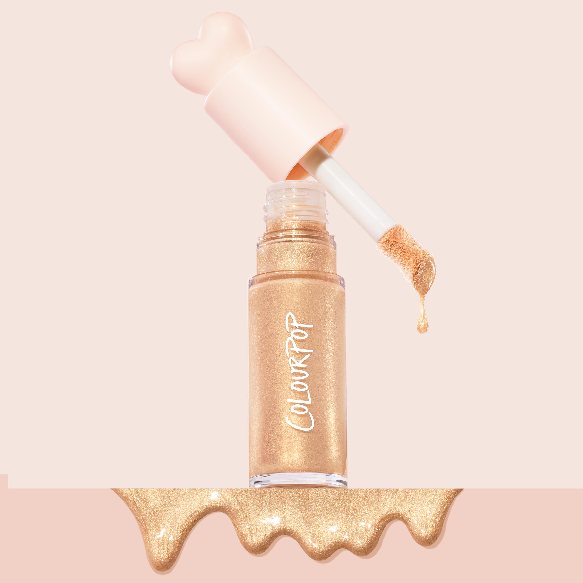 ColourPop Liquid Highlighter in Sugar Donut drips from a doe-foot applicator, a golden shimmer drop beside the clear tube with a pink heart-shaped cap, set on a soft blush backdrop with a glossy metallic swatch pooling at the bottom.