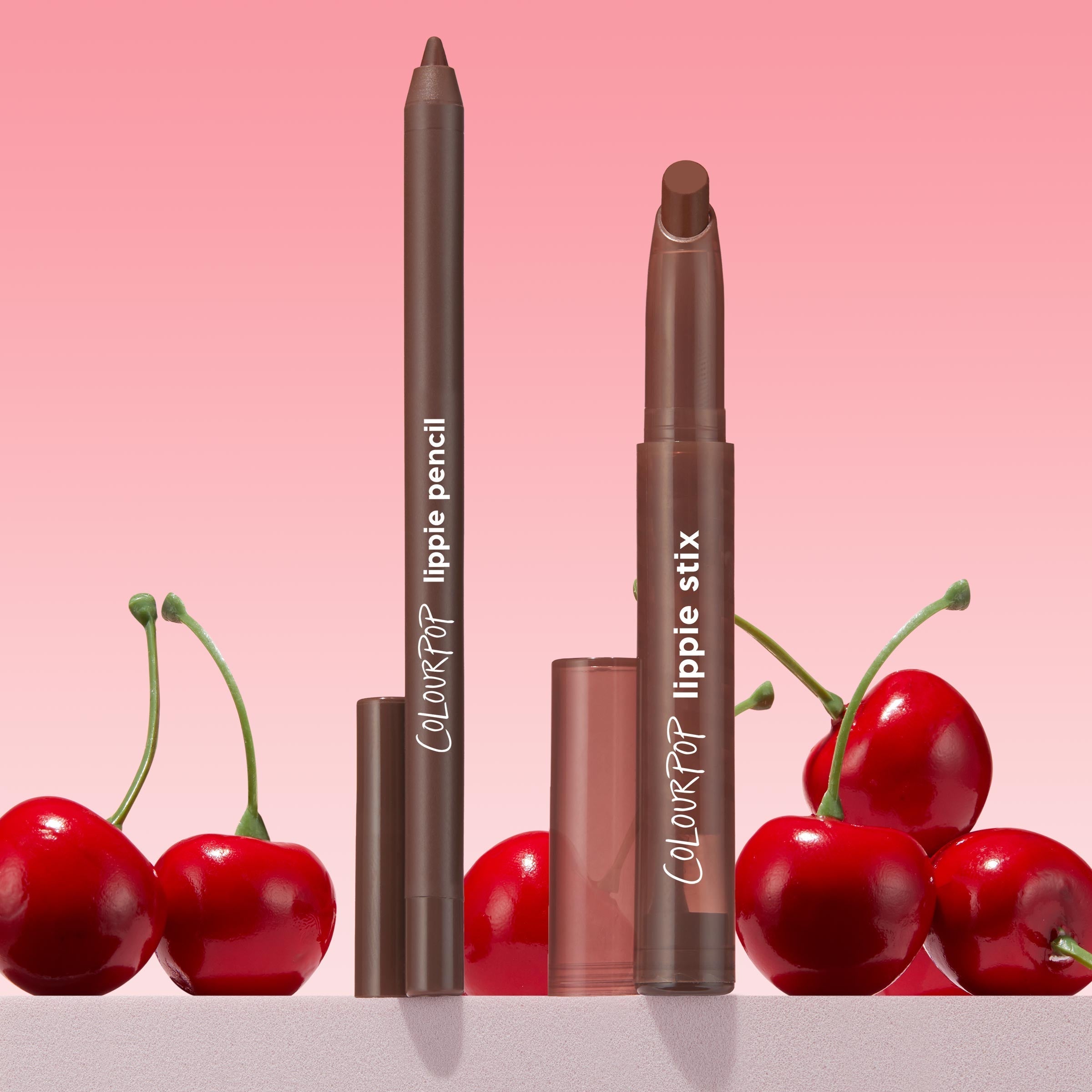 ColourPop Lippie Stix + Pencil Set, Lip Service, featuring a rich brown Lippie Stix and matching lip pencil standing uncapped with caps nearby, styled among glossy cherries against a pink gradient backdrop for a playful beauty product shot.