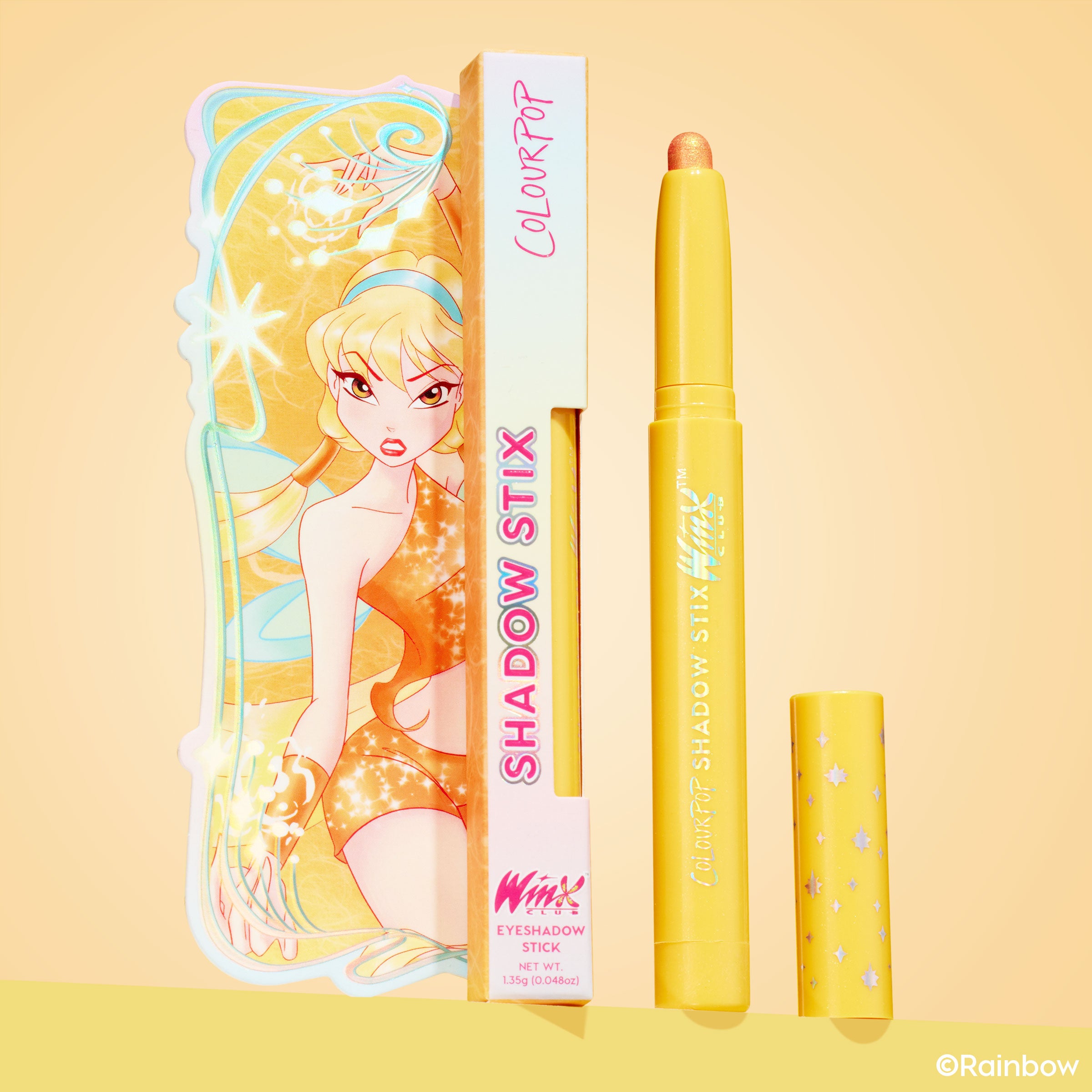 Rising Sun ColourPop Shadow Stix eyeshadow crayon stands uncapped to show its shimmery golden‑peach tip, next to a yellow star‑print cap and a Winx Club illustrated box on a warm peach background.