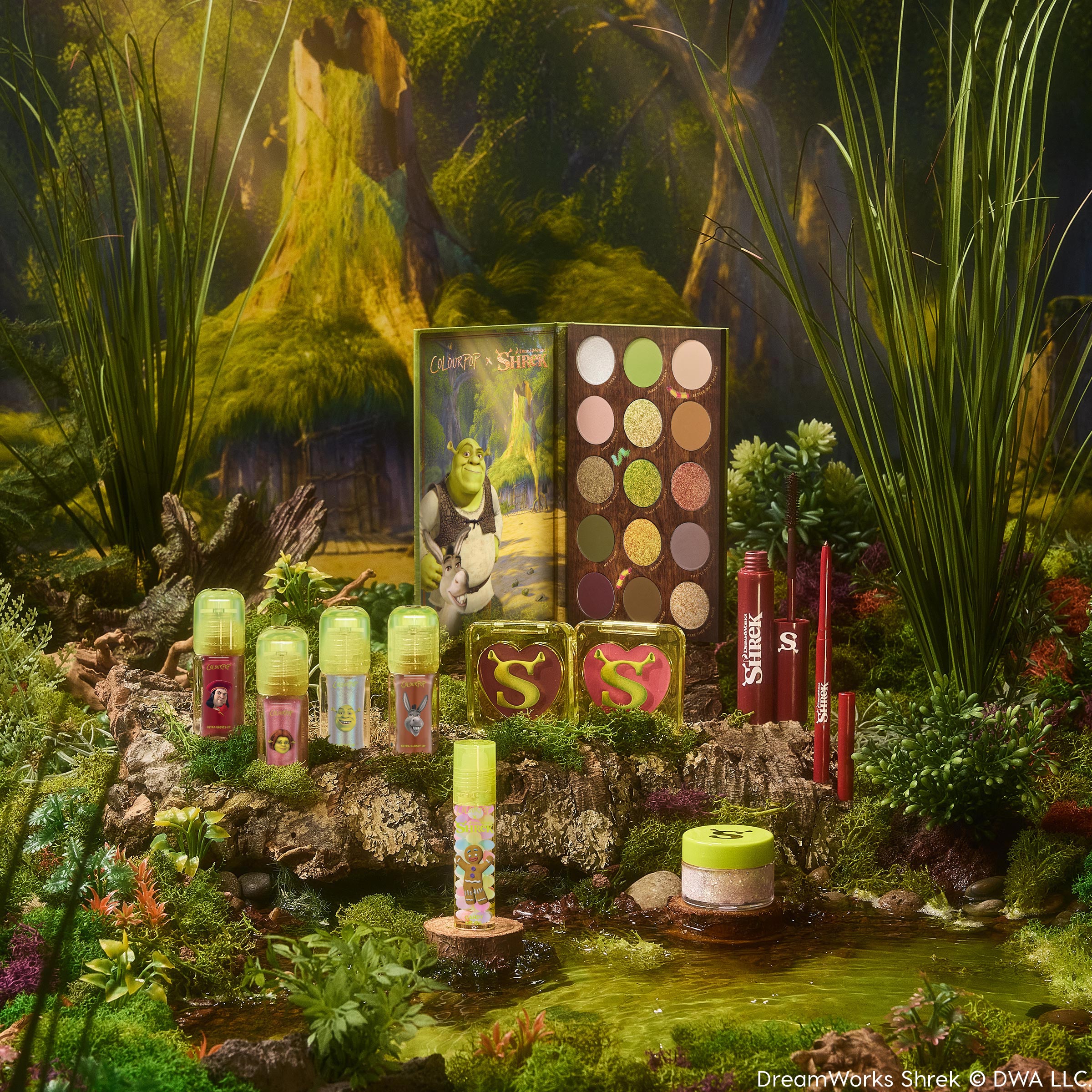 ColourPop X DreamWorks Shrek Full Collection Set showcasing an open 15-pan green-and-neutral eyeshadow palette, lip products, blush compacts with S logos, eyeliner, and highlighter, styled in a mossy swamp scene; ColourPop Full Collection Set.
