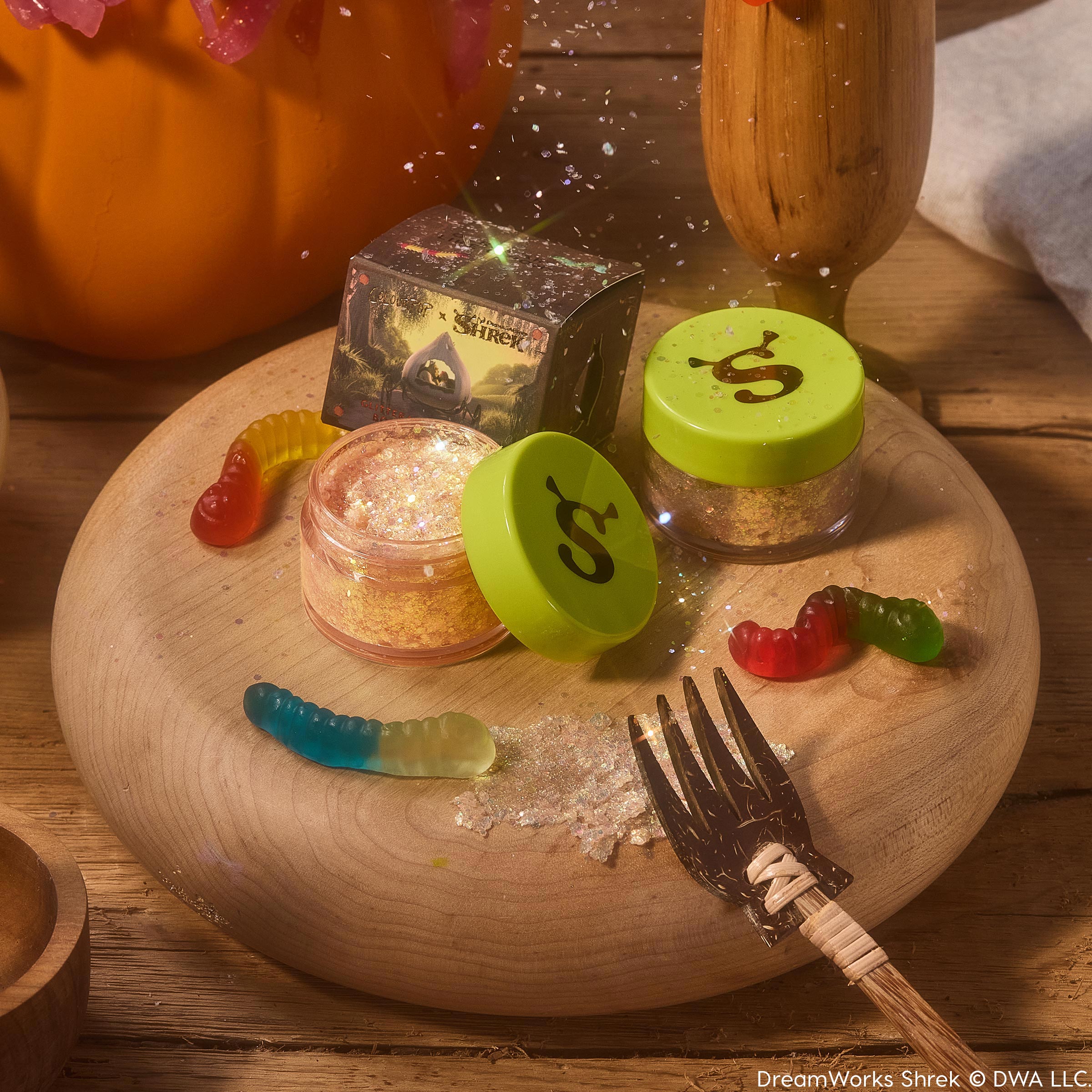 ColourPop Glitter Gel Ogres are Like Onions features an open jar of gold iridescent glitter gel with a lime green lid stamped with an ogre ear, beside a closed pot and Shrek x ColourPop box on a wooden board with gummy worms and scattered sparkles.