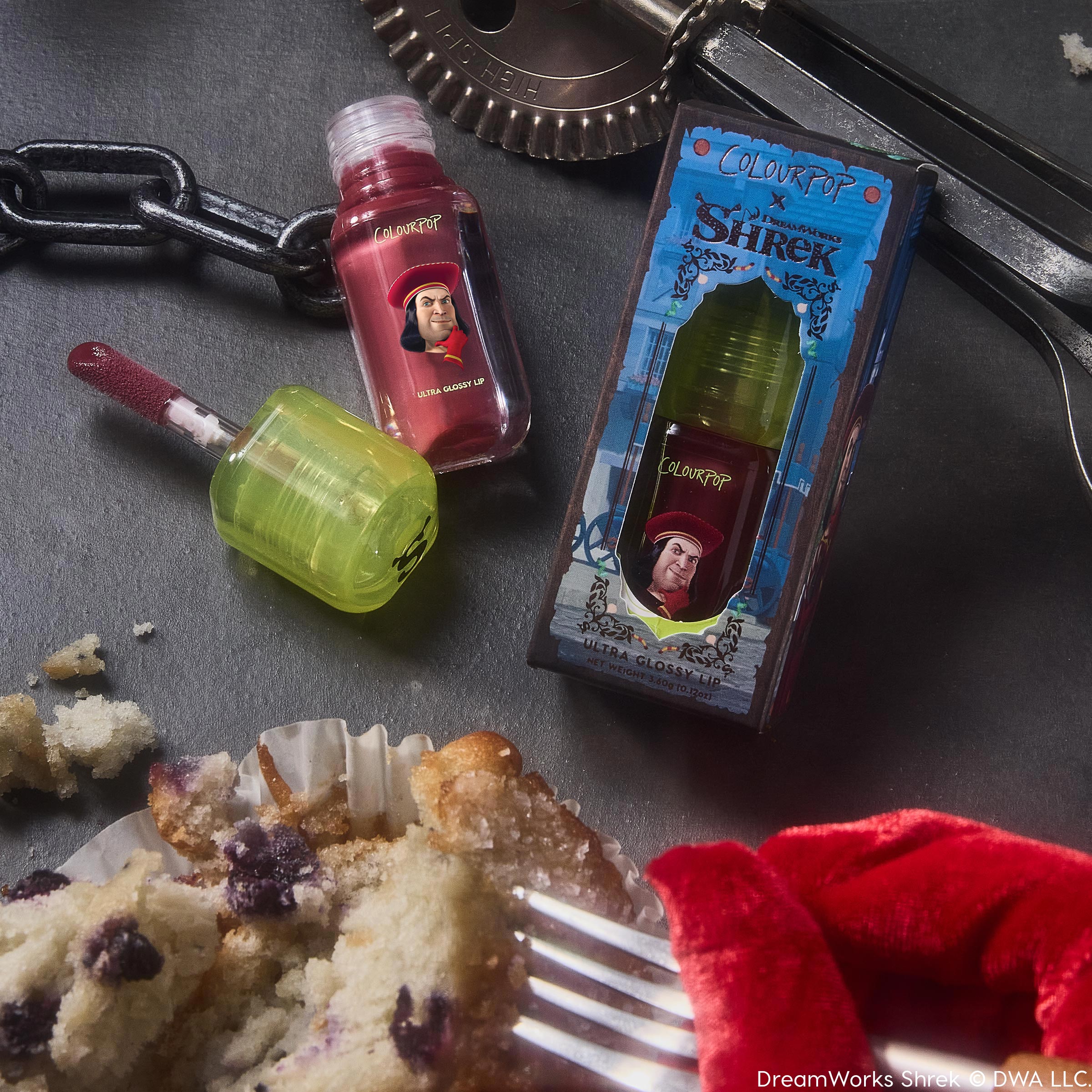 Short Lord ColourPop Ultra Glossy Lip - red tint with doe-foot applicator and neon green cap - rests open beside its Shrek-themed box featuring Lord Farquaad on a dark tabletop with crumbs, chain, and kitchen tools.