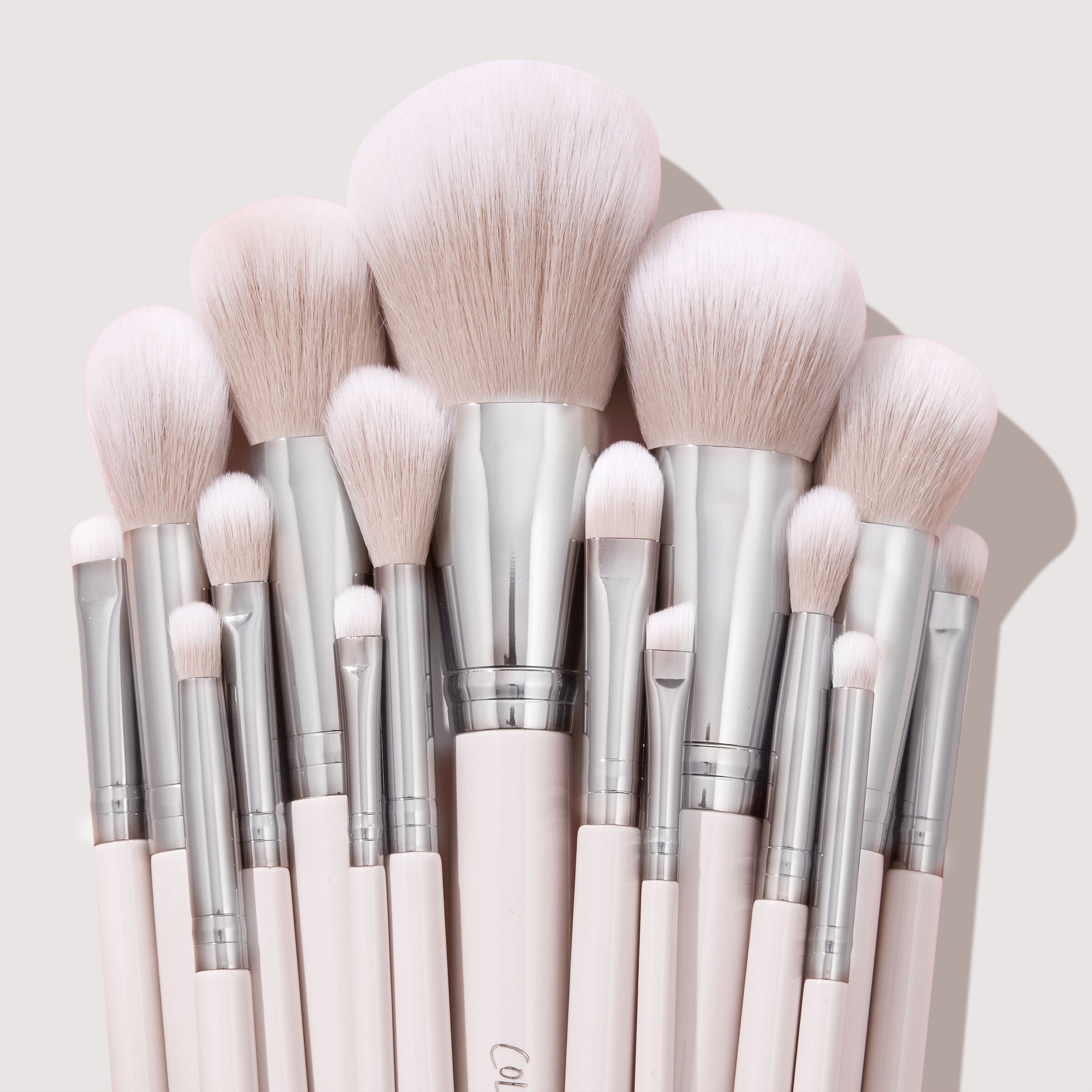 Stone Cold ColourPop Makeup Brush Kit fanned upright, displaying powder, blush, contour, and eye brushes with fluffy blush-pink bristles, silver ferrules, and pale pink handles, stacked tightly against a soft beige backdrop.