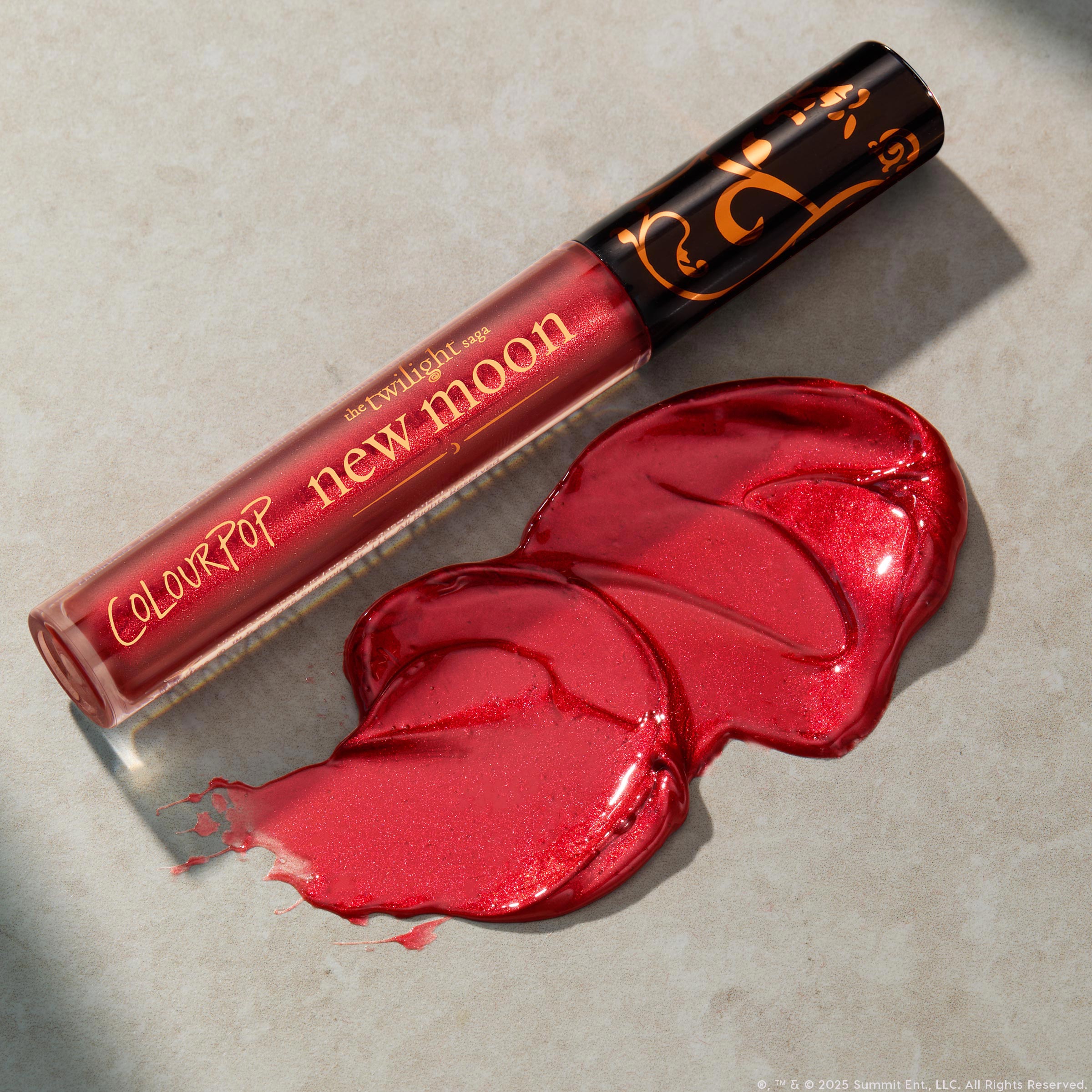 ColourPop Ultra Glossy Lip in Runs with Vampires lies diagonally with a glossy ruby-red, micro-shimmer swatch beside it; clear tube shows gold “new moon” lettering and a black cap with gold scrollwork on a neutral stone surface.