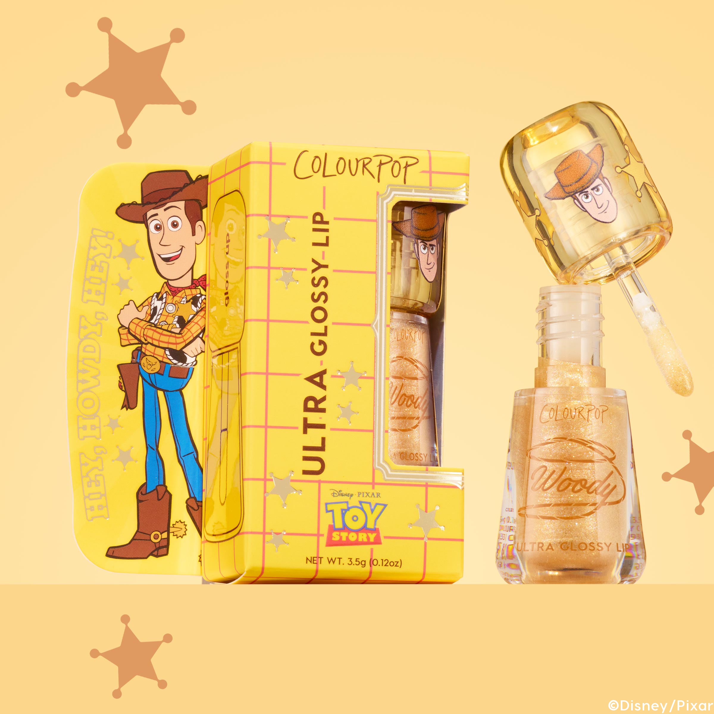 ColourPop Ultra Glossy Lip in Favorite Deputy shown open with the doe-foot applicator lifted, revealing a sheer gold sparkle gloss, the cap printed with Woody, beside a yellow Toy Story box on a warm yellow backdrop with sheriff stars.