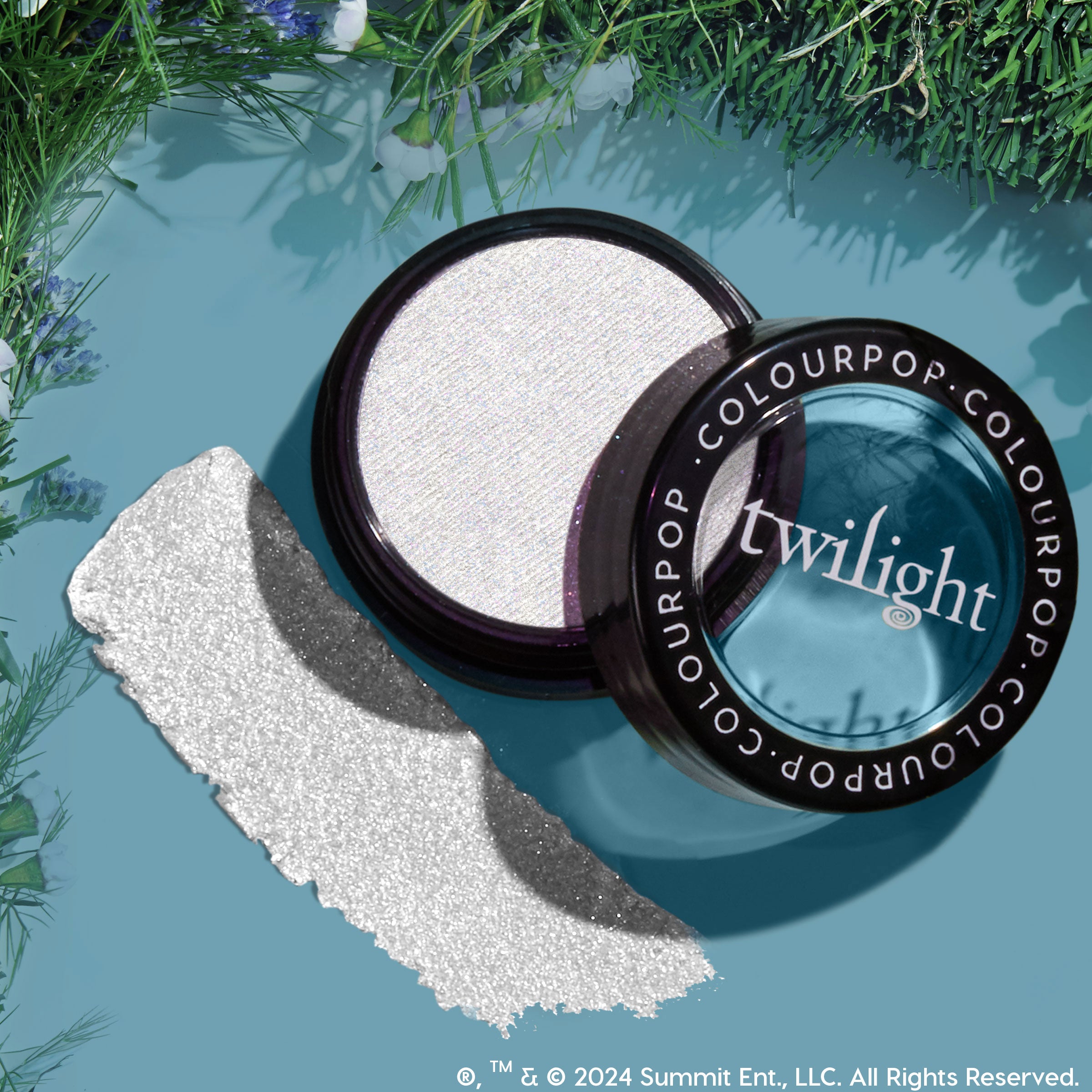 Vampire Skin ColourPop Super Shock Highlighter gleams in an open jar, showing an icy white, sparkly pan with the Twilight-branded lid and a bold swatch, styled as a flat lay on a teal backdrop bordered with green foliage.