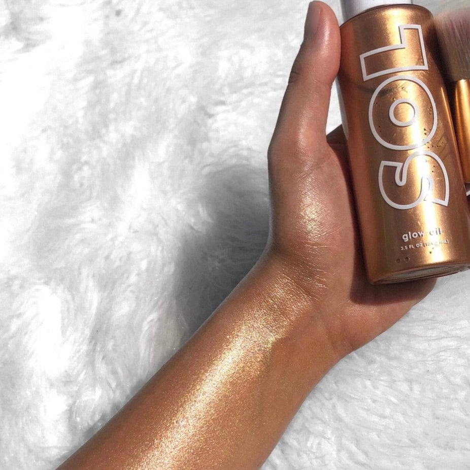 Warm Gold SOL Glow Oil bottle from ColourPop SOL Glow Oil held in a hand, showing a shimmery warm gold swatch on the forearm; metallic bronze bottle with white SOL logo, 3.5 fl oz, beside a makeup brush on a white faux-fur background.