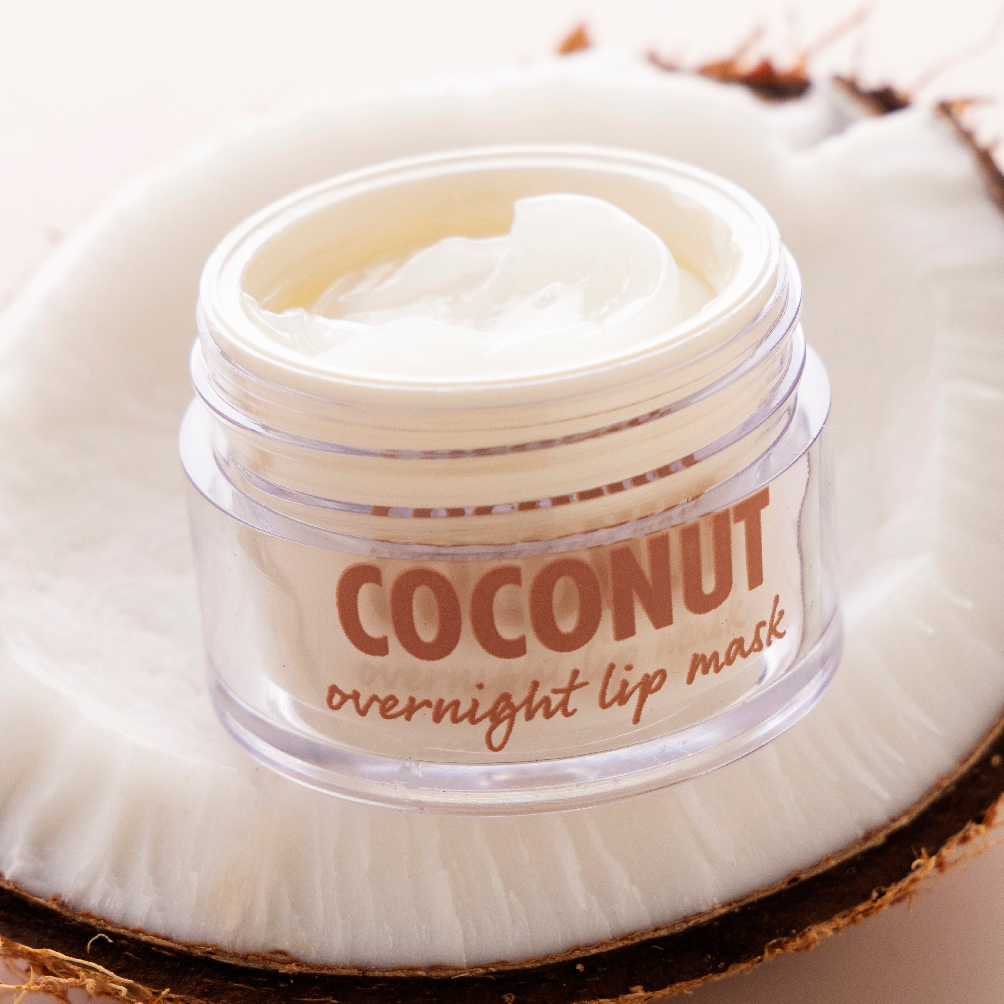 Open jar of Coconut overnight lip mask showing a swirl of creamy balm, resting on a halved coconut; clear pot with tan lettering highlights hydrating formula for dry lips; ColourPop Lip Mask product shot.