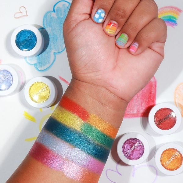 Pug Life Super Shock Shadow swatched on a forearm, showing smooth, metallic payoff, surrounded by open ColourPop Super Shock Shadow pots in rainbow hues; hand with rainbow nail art rests over a doodled cloud and rainbow background.
