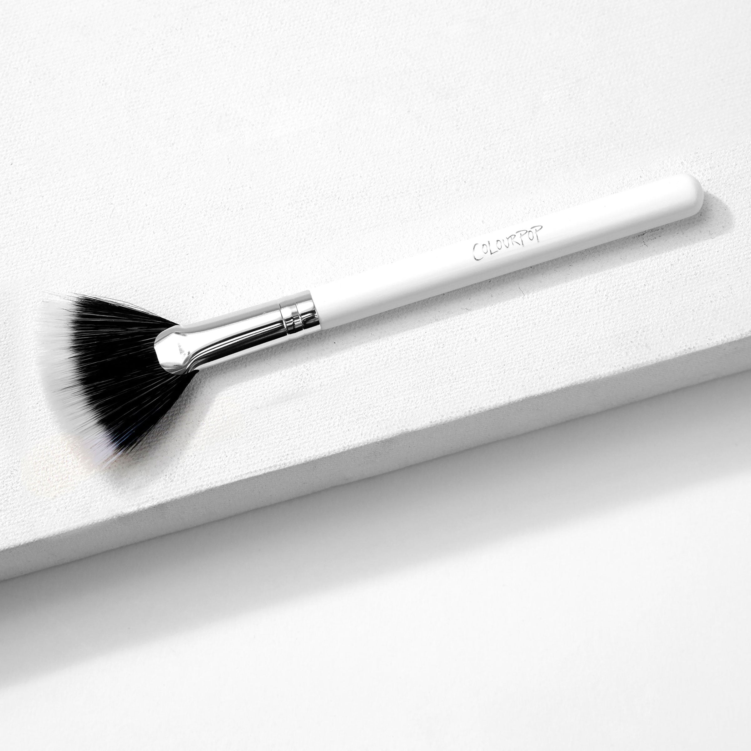ColourPop Fan Brush makeup brush resting diagonally on a white textured block, featuring duo-fiber black-to-white fanned bristles, a silver ferrule, and a white handle embossed with ColourPop, designed for sweeping highlighter.
