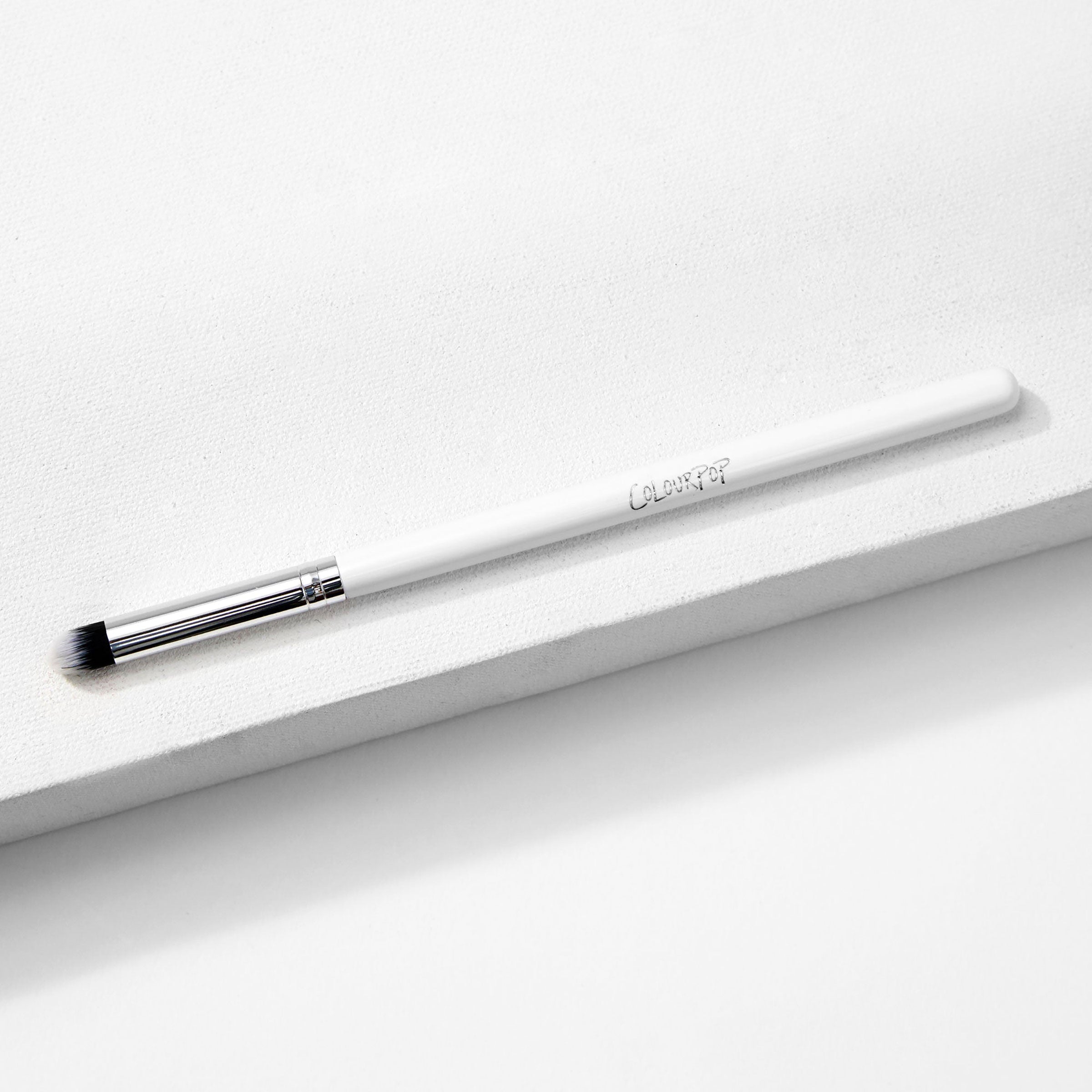 ColourPop Pencil Brush resting diagonally on a white textured block, a makeup brush with a slim white handle and logo, silver ferrule, and small tapered black-and-white bristles for precise eyeshadow and eyeliner smudging.
