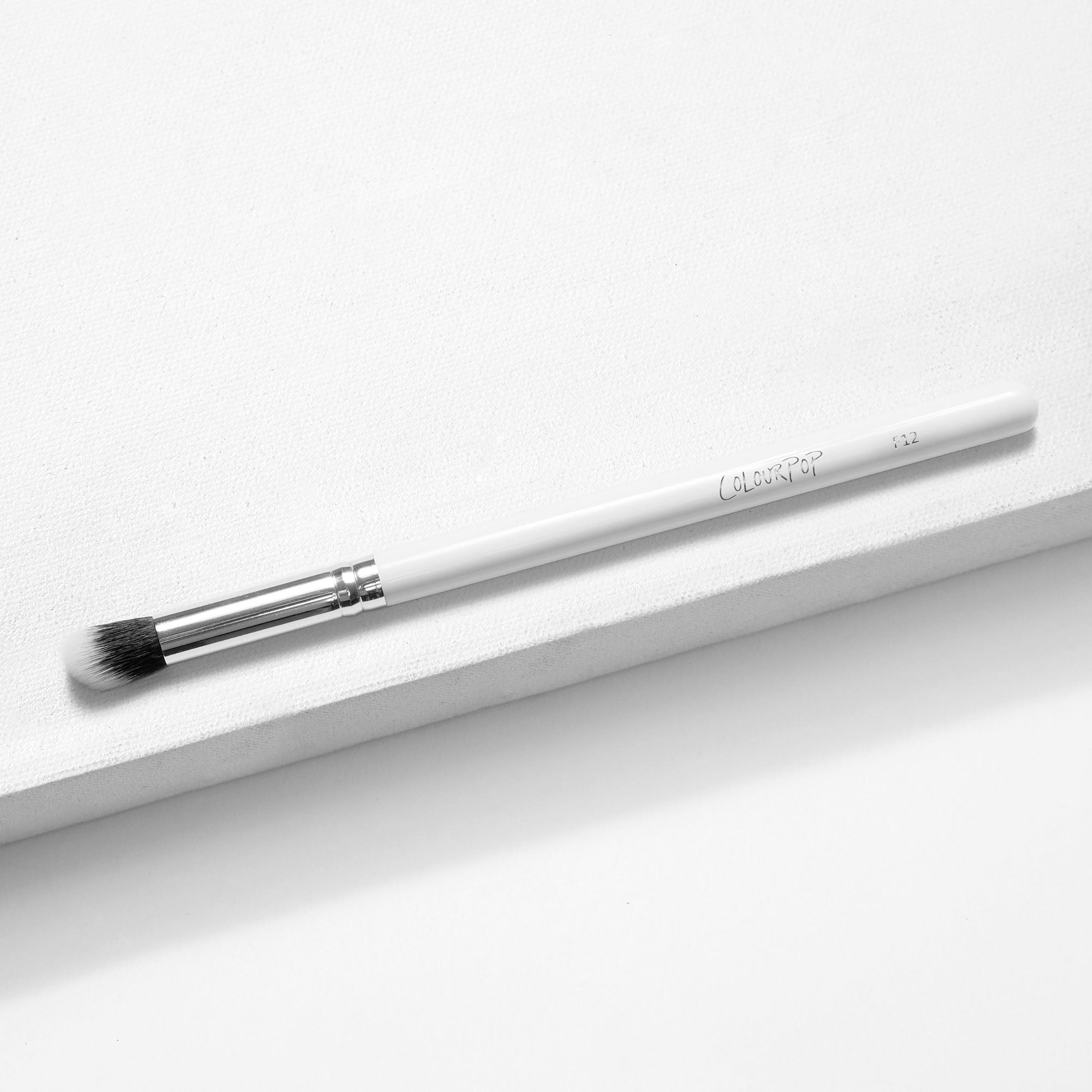 ColourPop Small Detail Brush lying diagonally on a white textured backdrop, showcasing tapered black-to-white synthetic bristles, a silver ferrule, and a slim white handle; precision makeup brush for detailed eyeshadow and smudging.