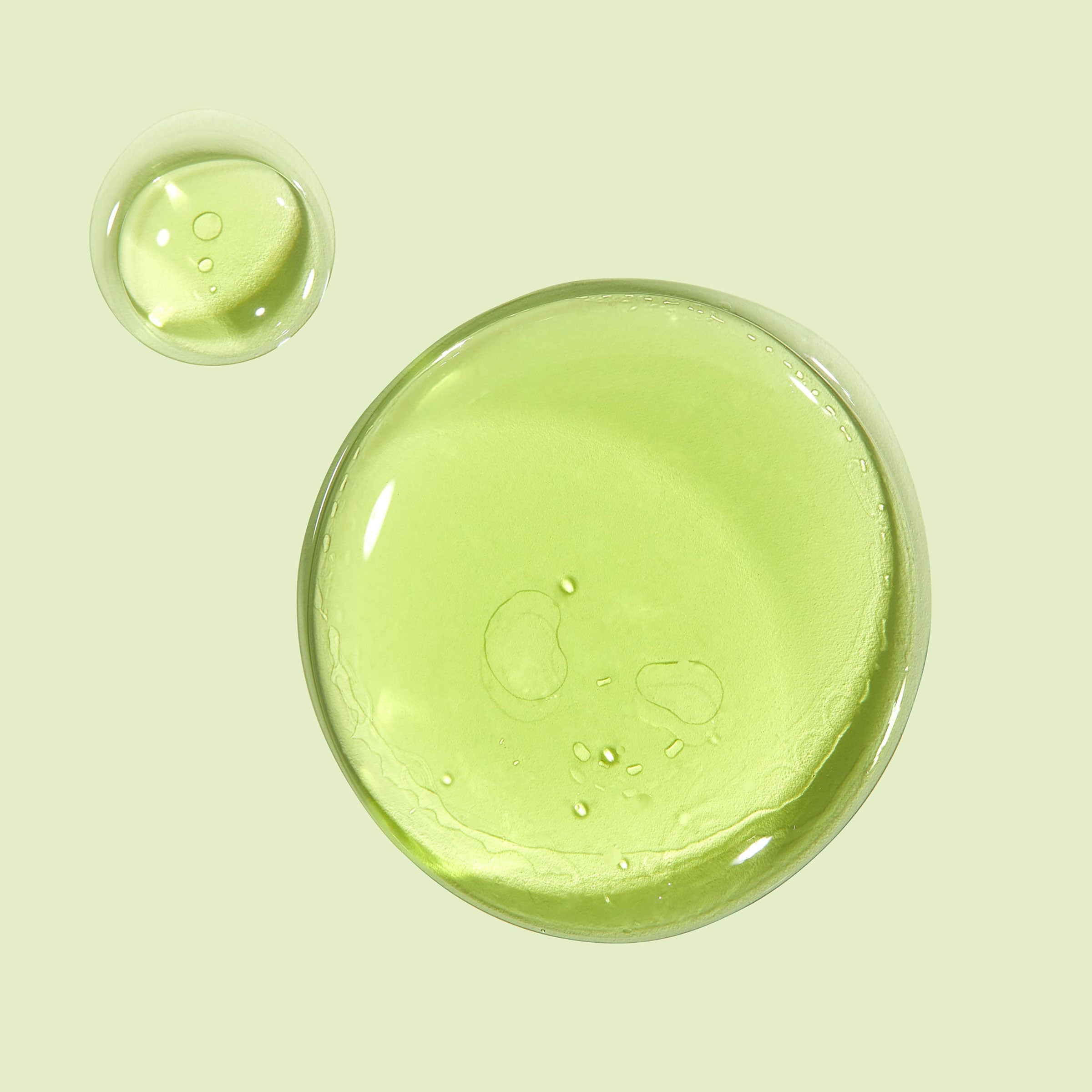Avocado + Aloe Double Shot face serum forming glossy, translucent green drops that pool on a pale mint background, highlighting a lightweight, hydrating texture; ColourPop Face Serum.